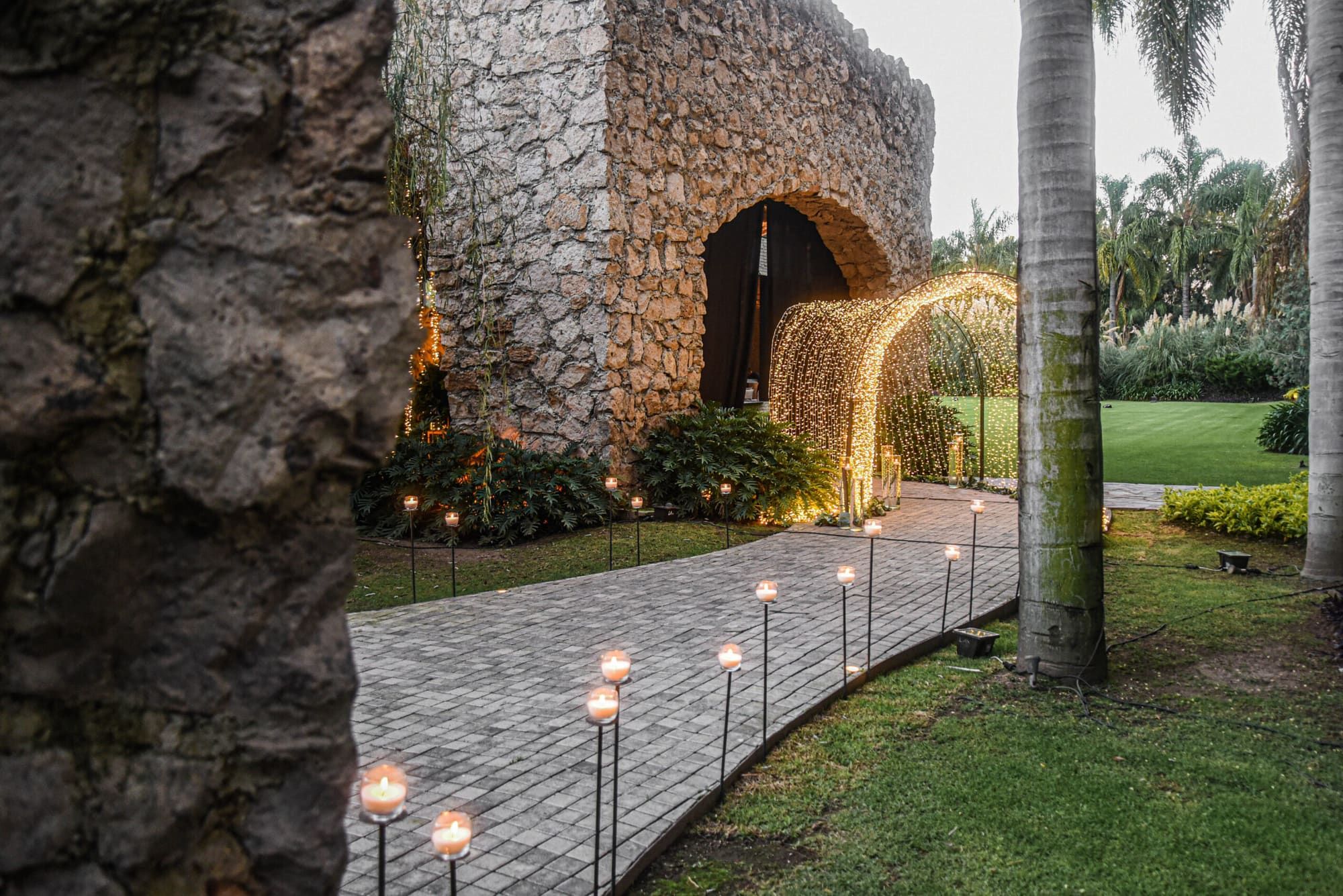 Arco de piedra con túnel de luces cálidas al final de un camino empedrado bordeado de velas