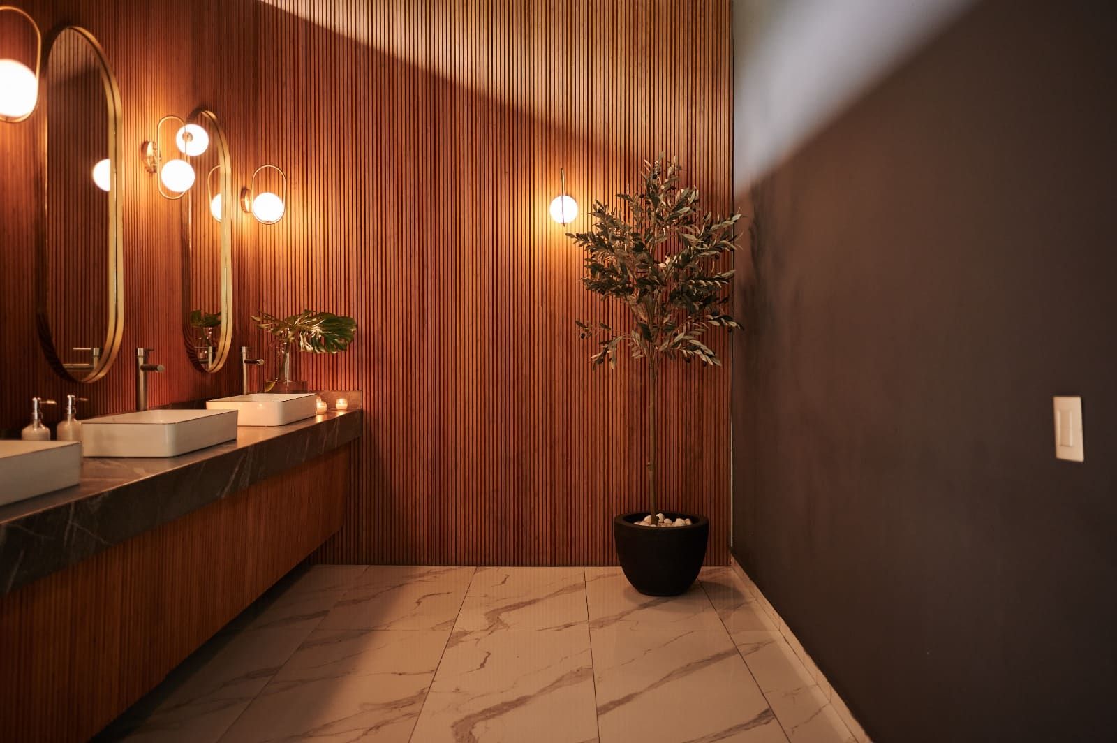 Baño con muro de madera, lavabos sobre encimera oscura, espejos ovalados y una planta en maceta negra