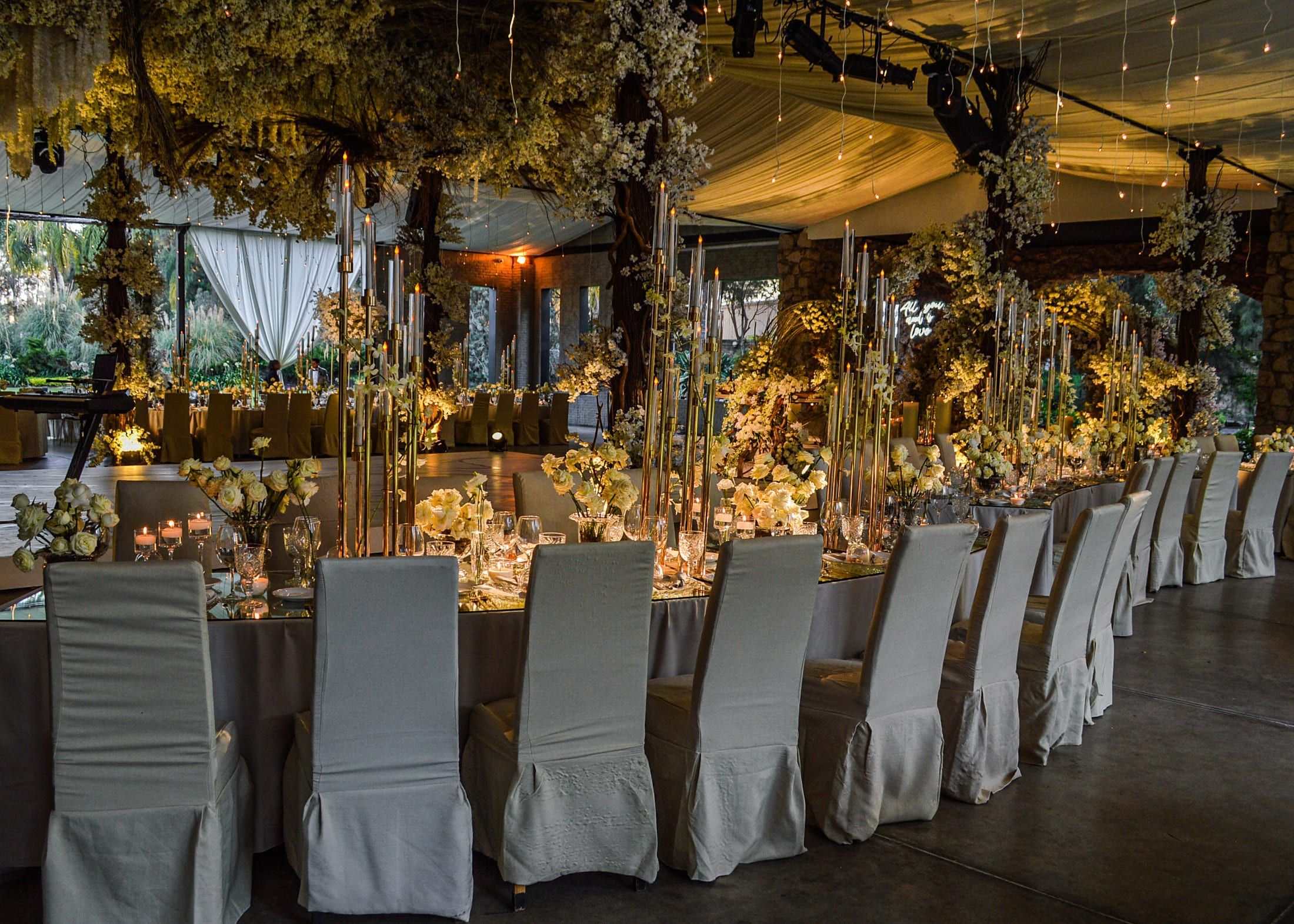 Larga mesa de banquete con sillas vestidas en tono claro y flores a lo largo, bajo un techo floral