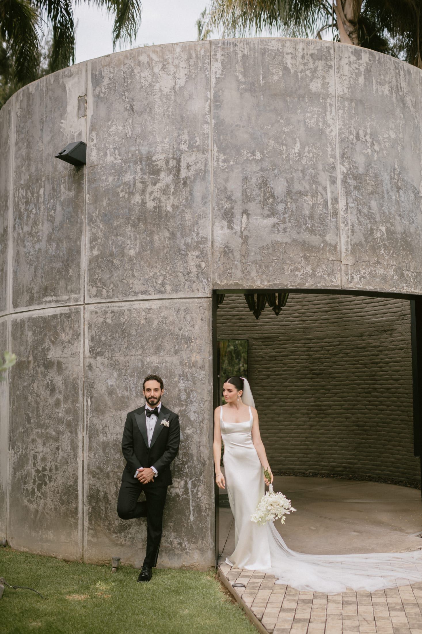 Los novios junto al cilindro de concreto, él recargado en el muro y ella con vestido blanco y ramo
