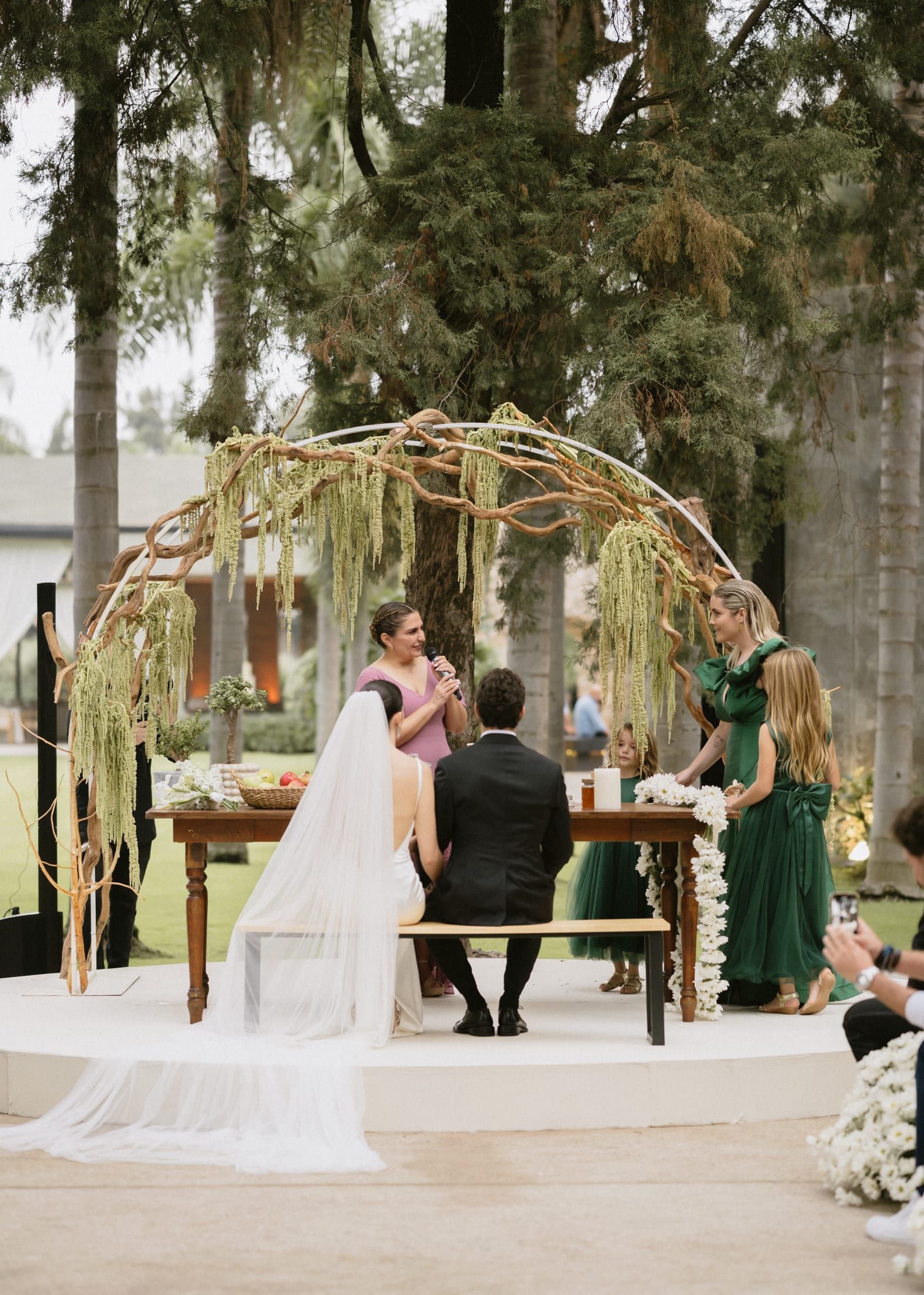 Ceremonia bajo un arco de ramas y follaje, con la pareja sentada frente a una mesa de madera