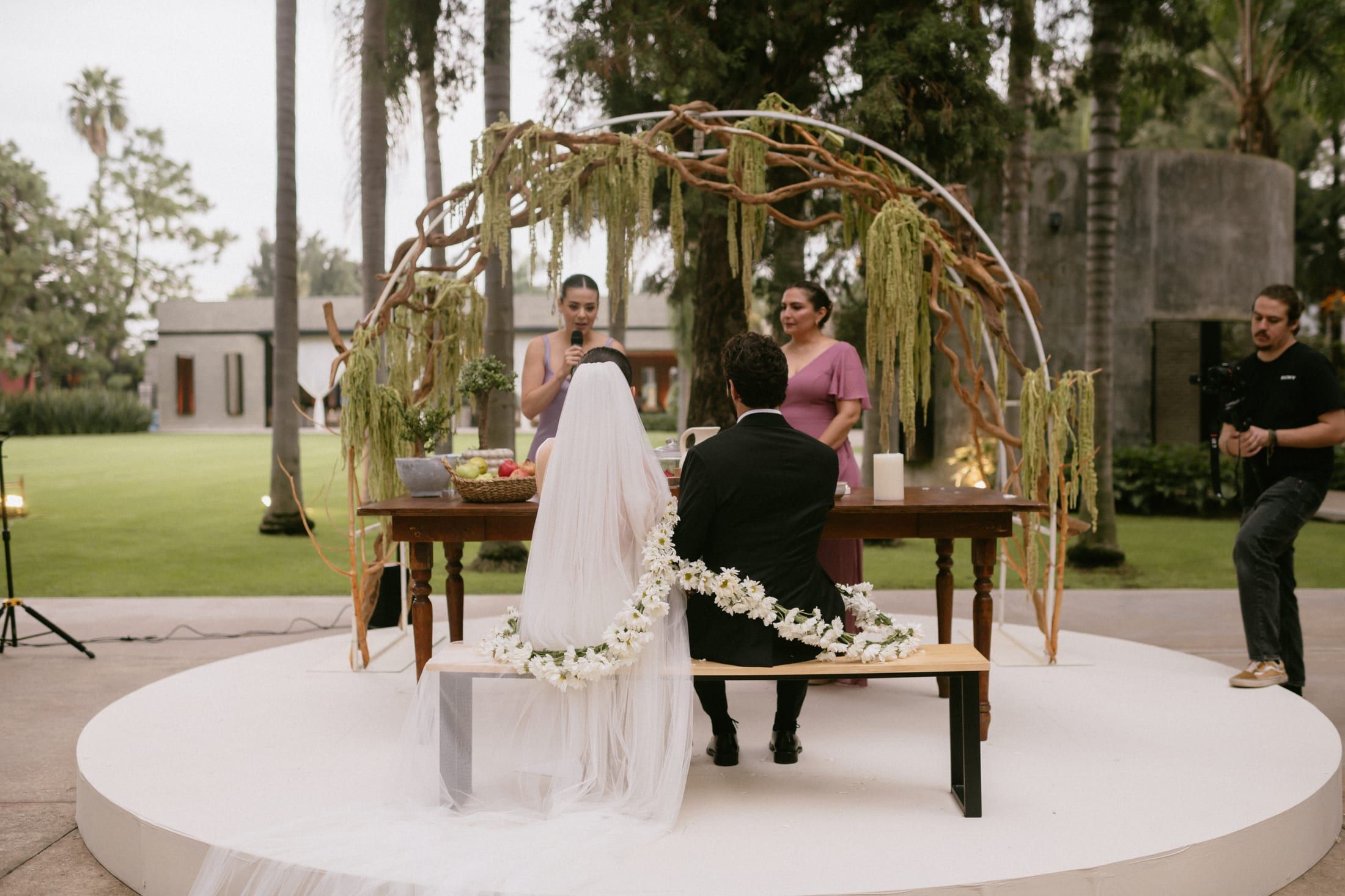 Ceremonia sobre una tarima blanca en el jardín, con arco de ramas y una guirnalda de flores sobre el banco
