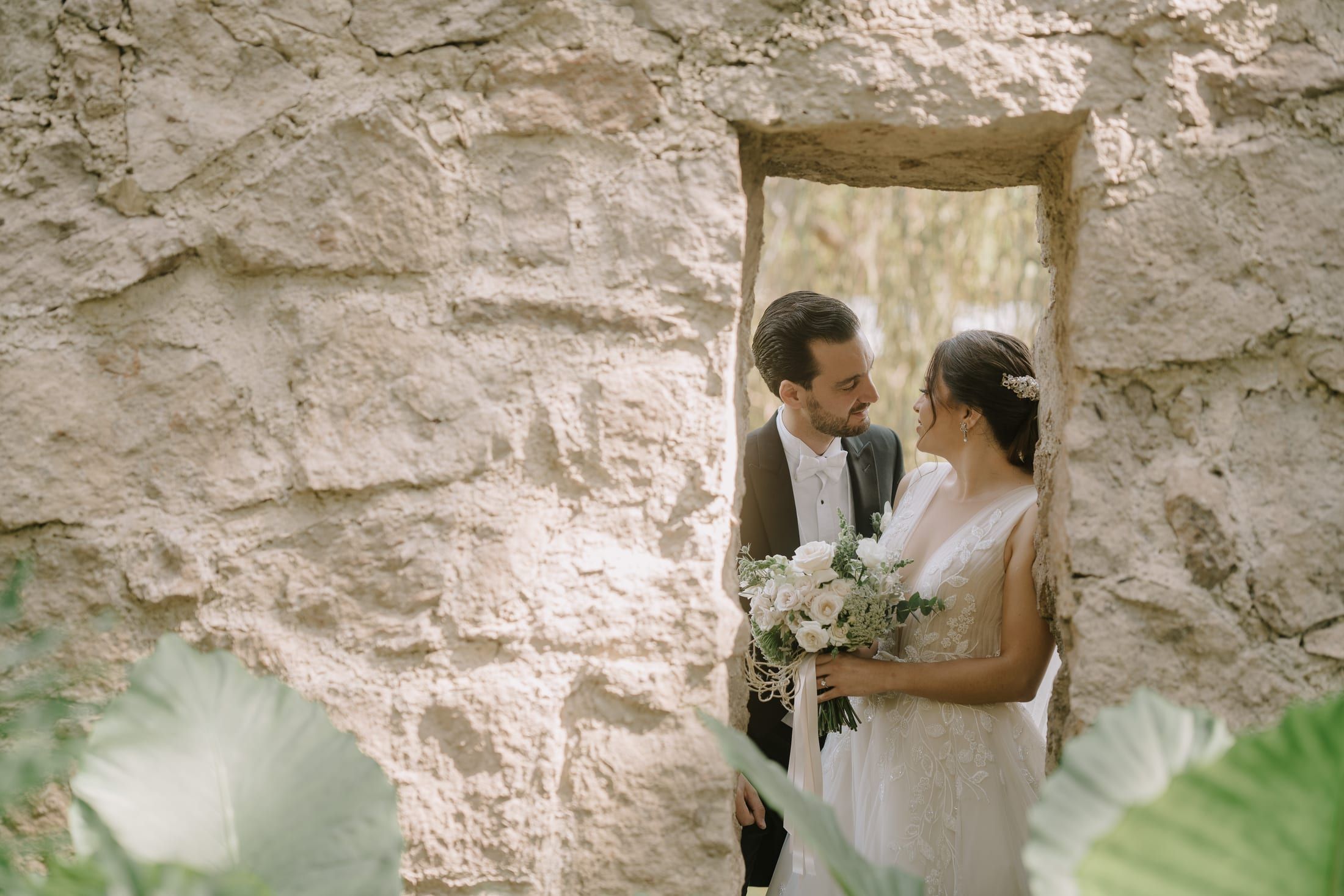 Los novios frente a frente, asomados por una ventana de piedra entre hojas grandes
