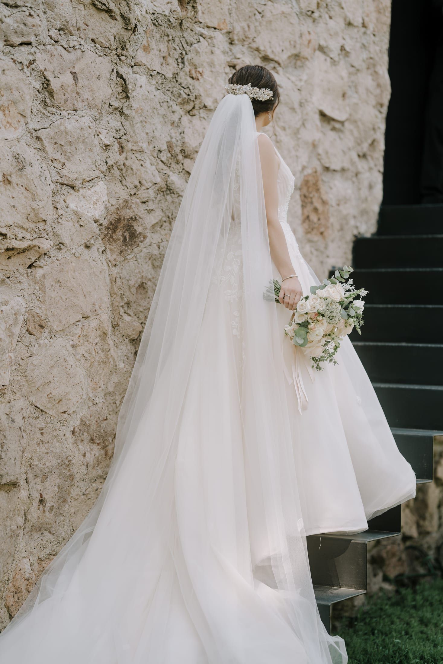 La novia de espaldas con velo largo y ramo, junto a un muro de piedra