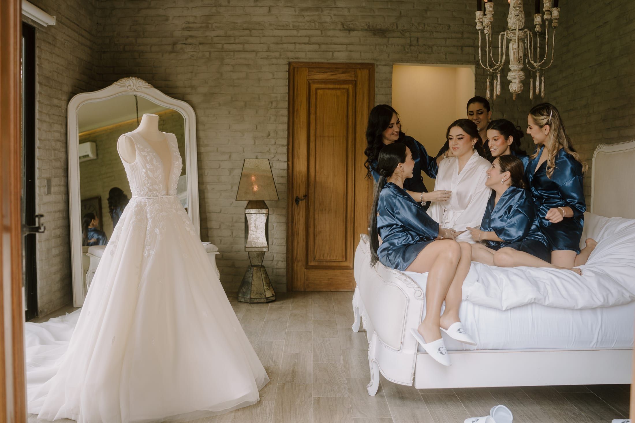 Damas con batas azules sentadas en la cama de la suite, junto al vestido de novia en el maniquí