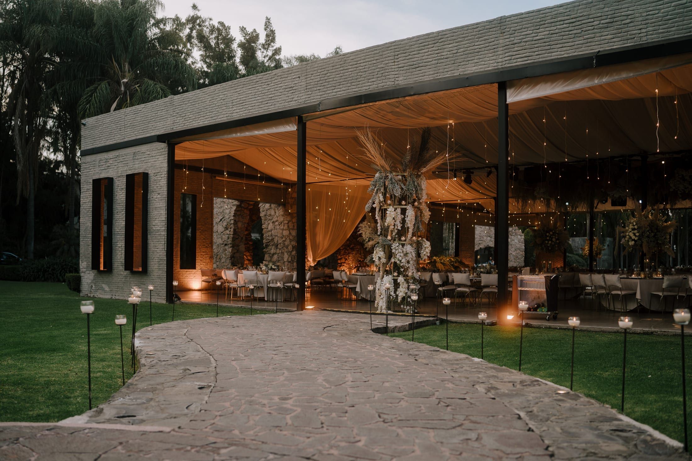 Fachada del salón al atardecer con un camino de velas y la recepción iluminada al fondo
