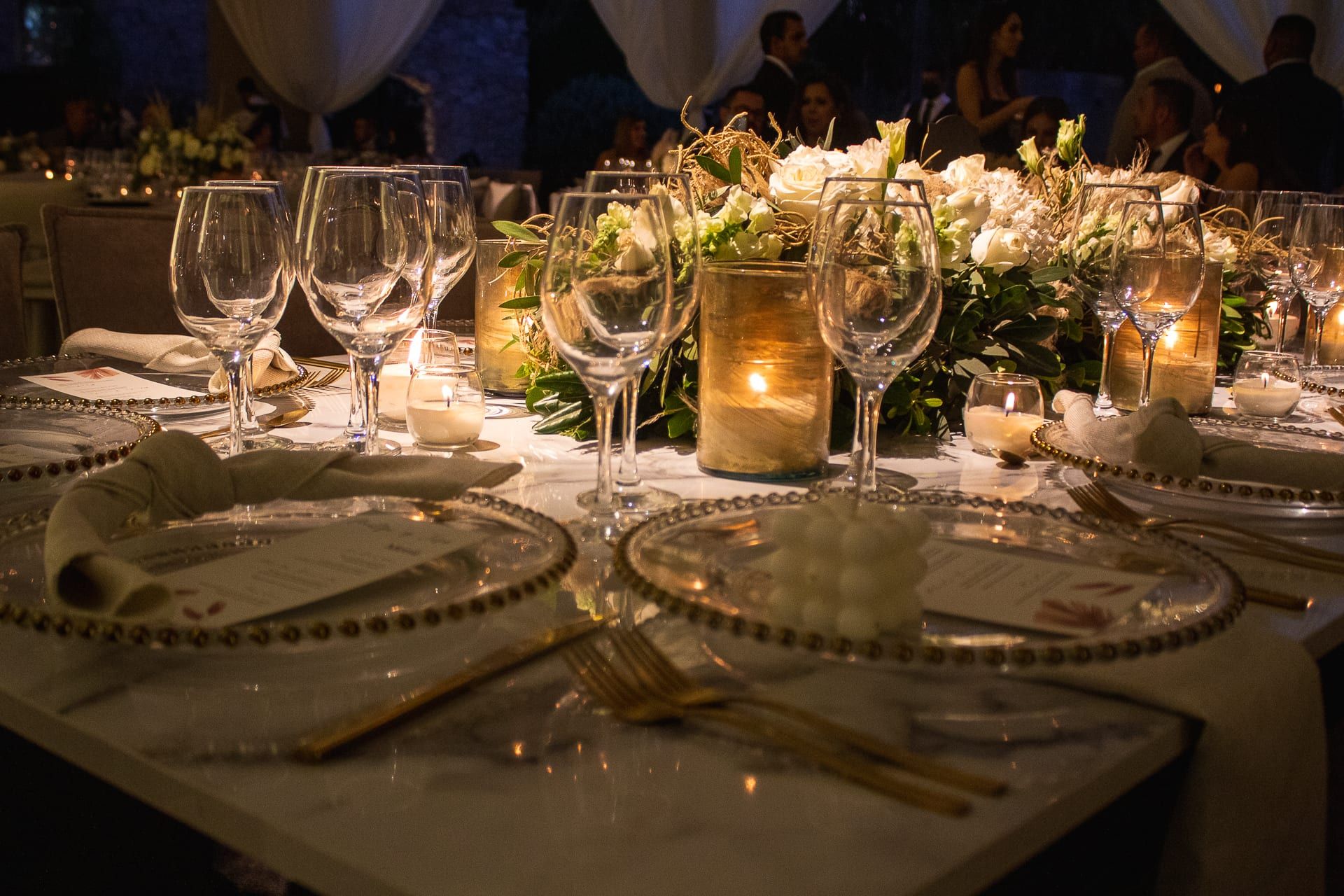 Mesa con vajilla de cuentas doradas, copas, velas y centros de flores blancas