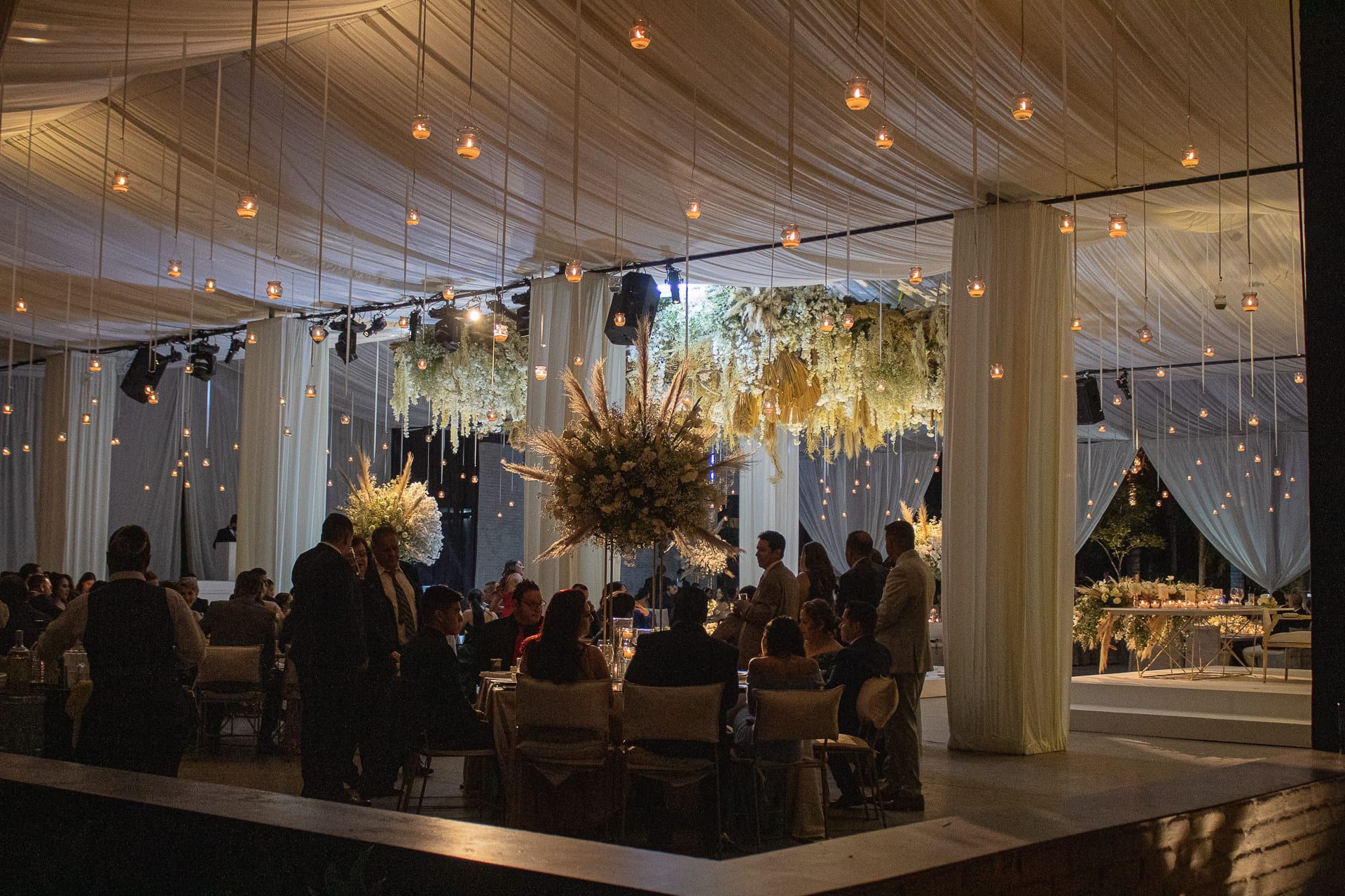Vista del salón desde el exterior, con cortinas, luces colgantes y los invitados sentados