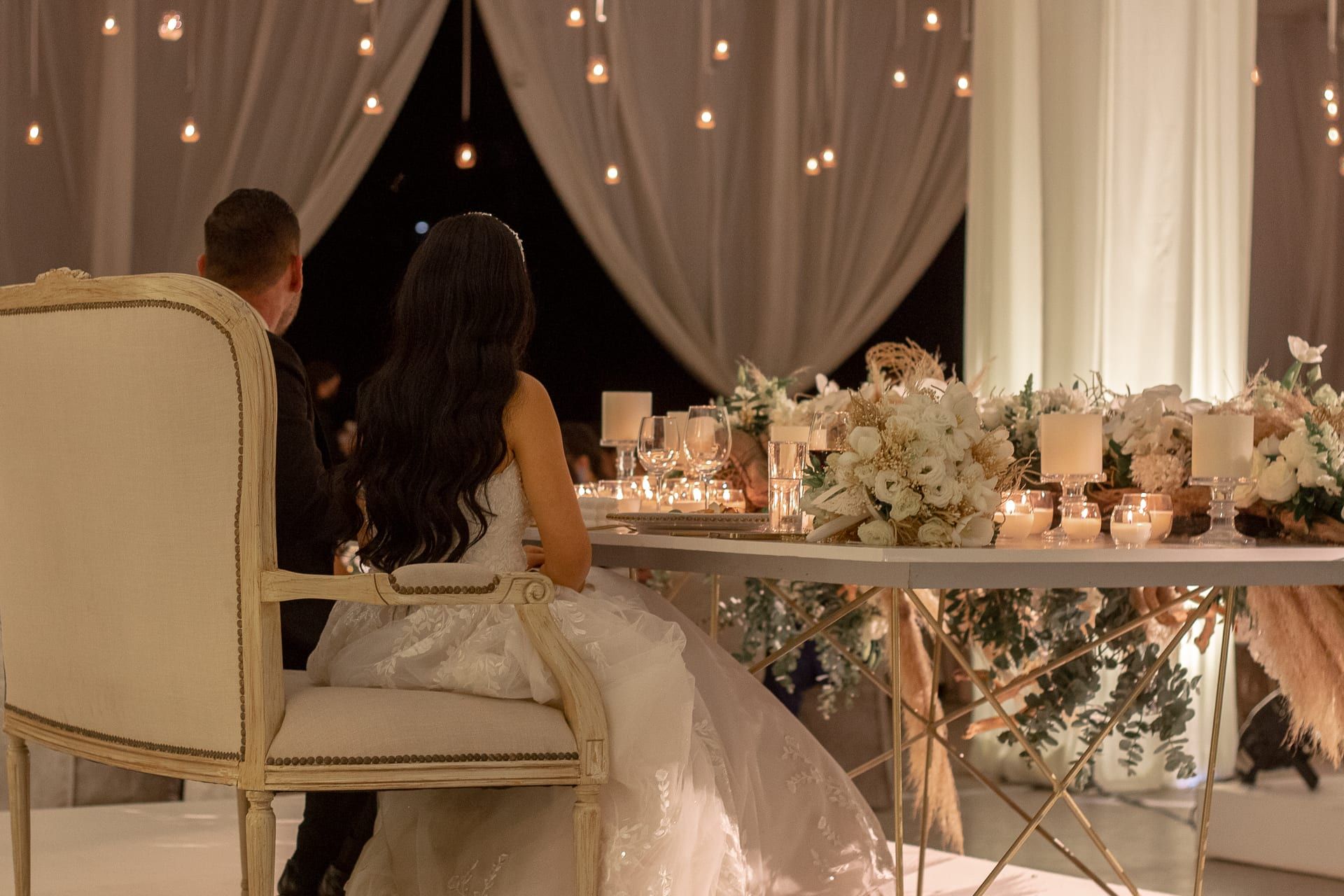 Los novios de espaldas en su mesa, con flores y velas, frente a las cortinas