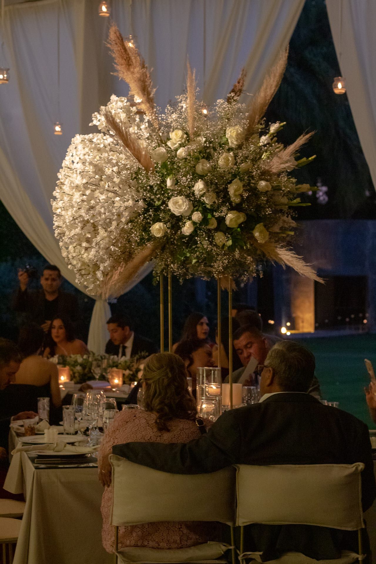 Arreglo alto de flores blancas y pampas junto a una mesa con invitados