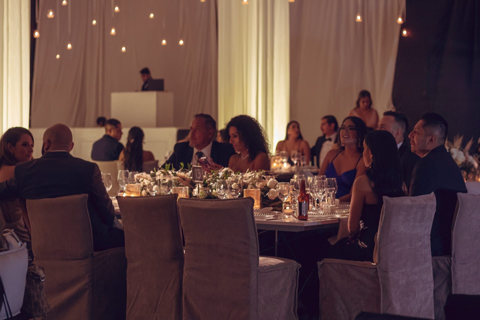 Invitados en mesas redondas bajo luces cálidas y cortinas de tela
