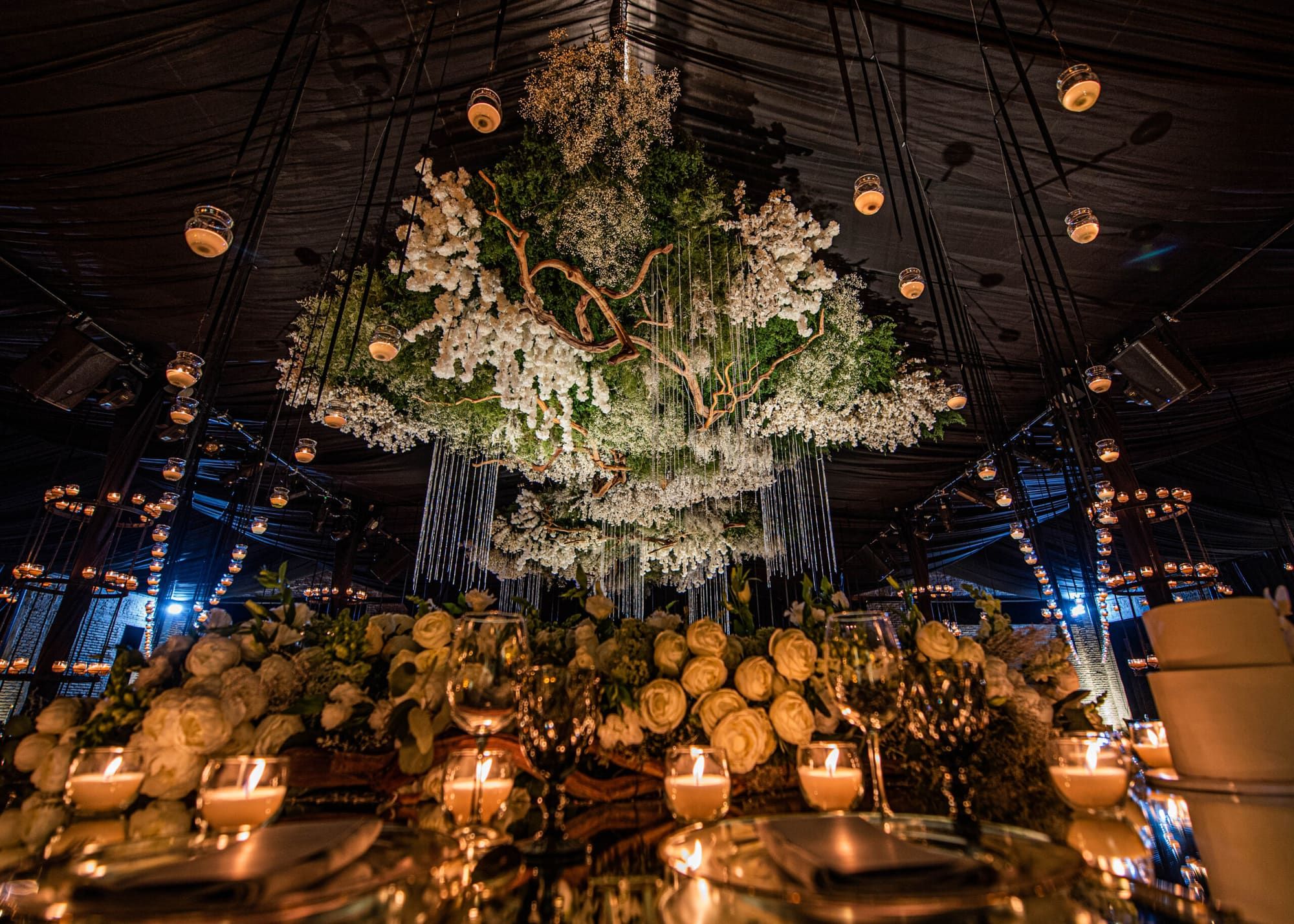 Instalación floral blanca con ramas colgando del techo oscuro sobre una mesa con rosas claras y velas
