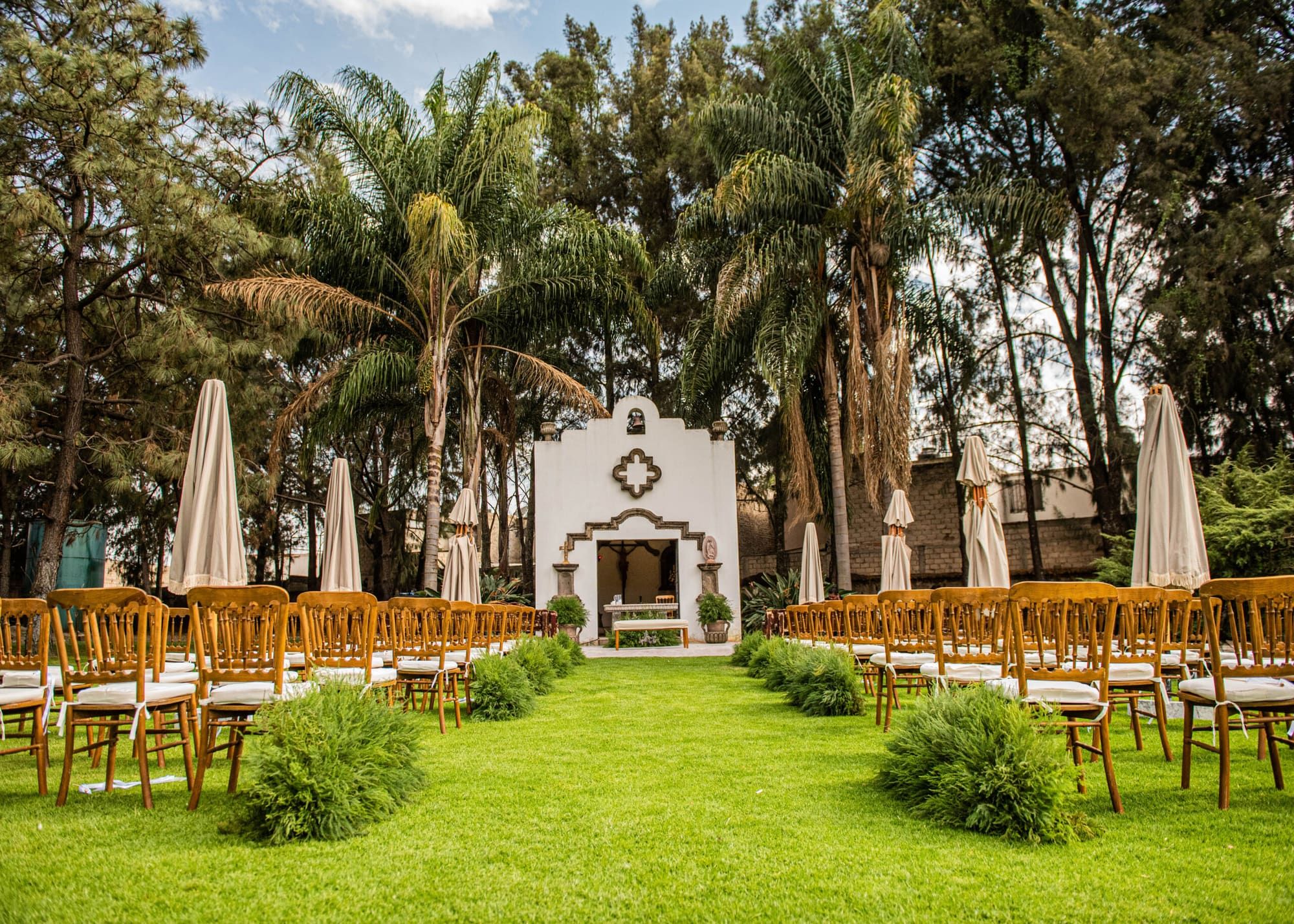 Capilla blanca al fondo de un jardín, con sillas de madera dispuestas entre palmeras y sombrillas