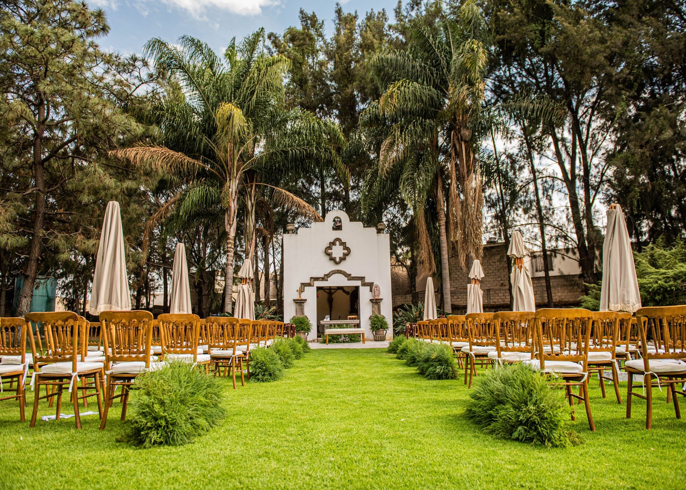 Capilla blanca al fondo de un pasillo de césped con filas de sillas de madera y sombrillas, entre palmeras