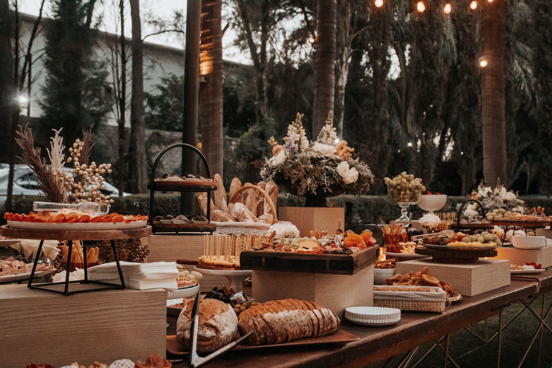 Buffet al aire libre con panes, charolas y arreglos florales, bajo palmeras y una línea de focos