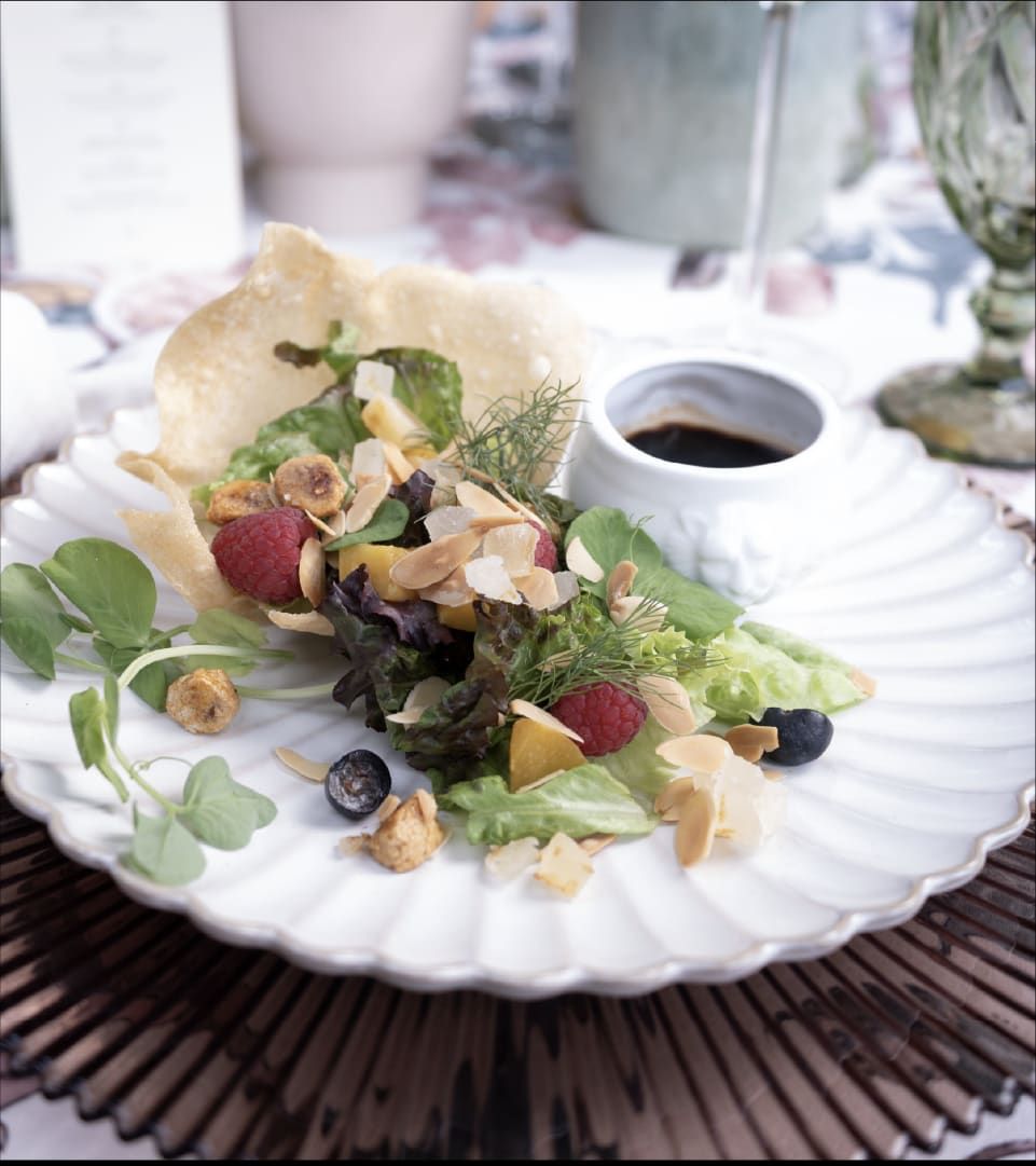 Ensalada de hojas tiernas con frutos rojos y crujientes sobre plato blanco, con un vasito de salsa oscura