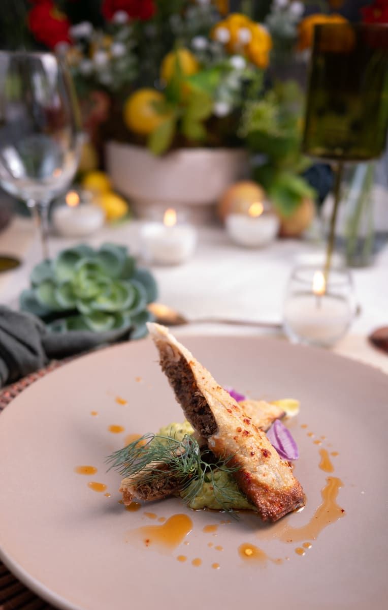 Platillo de piel crujiente sobre plato rosado, con flores de colores y velas desenfocadas al fondo