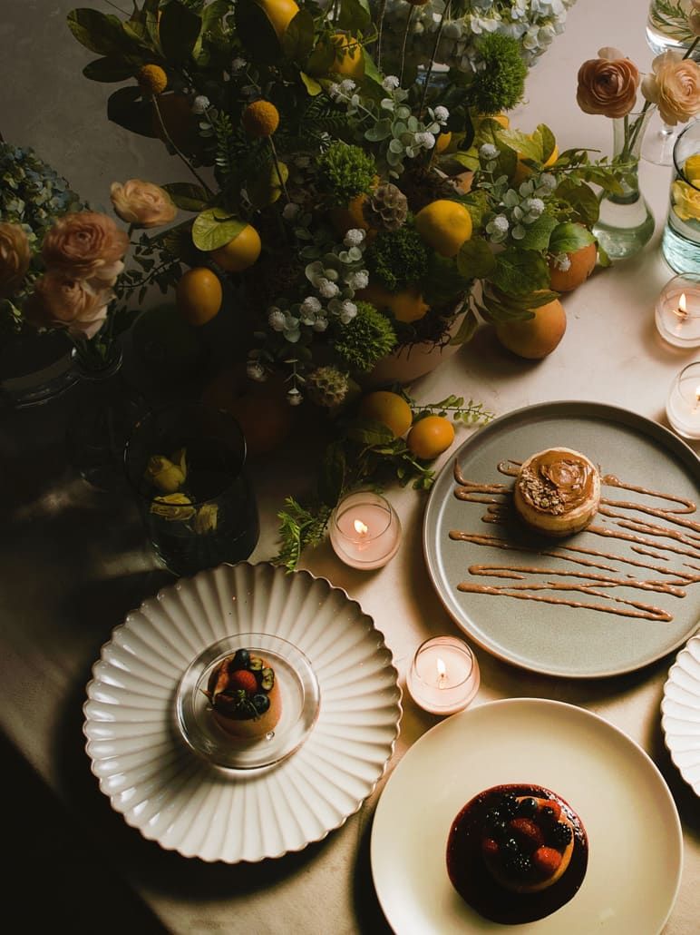 Postres sobre platos de cerámica junto a un arreglo floral con cítricos y velas pequeñas encendidas
