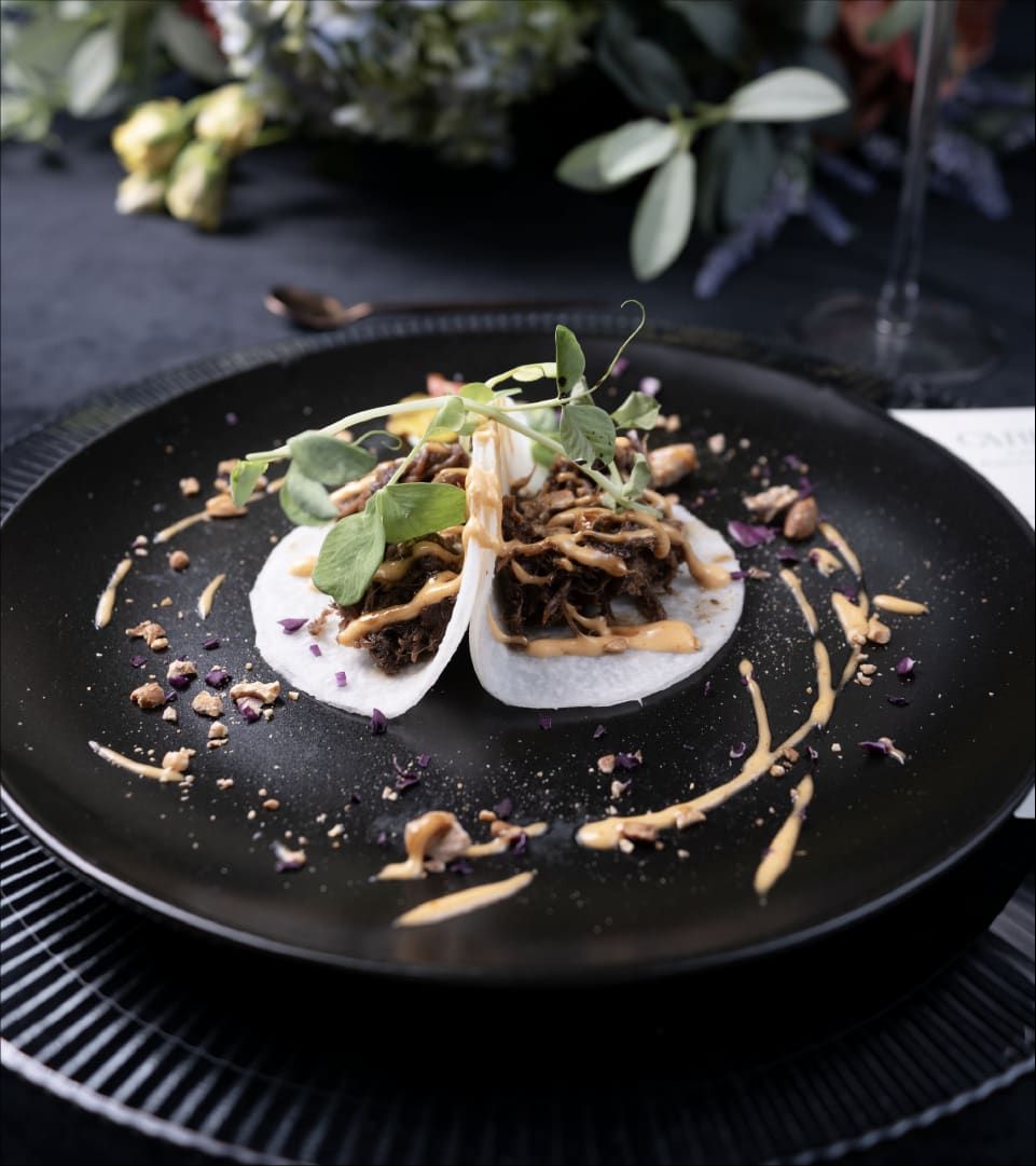 Dos tacos en tortilla blanca con carne deshebrada y hierbas sobre un plato negro con salsa