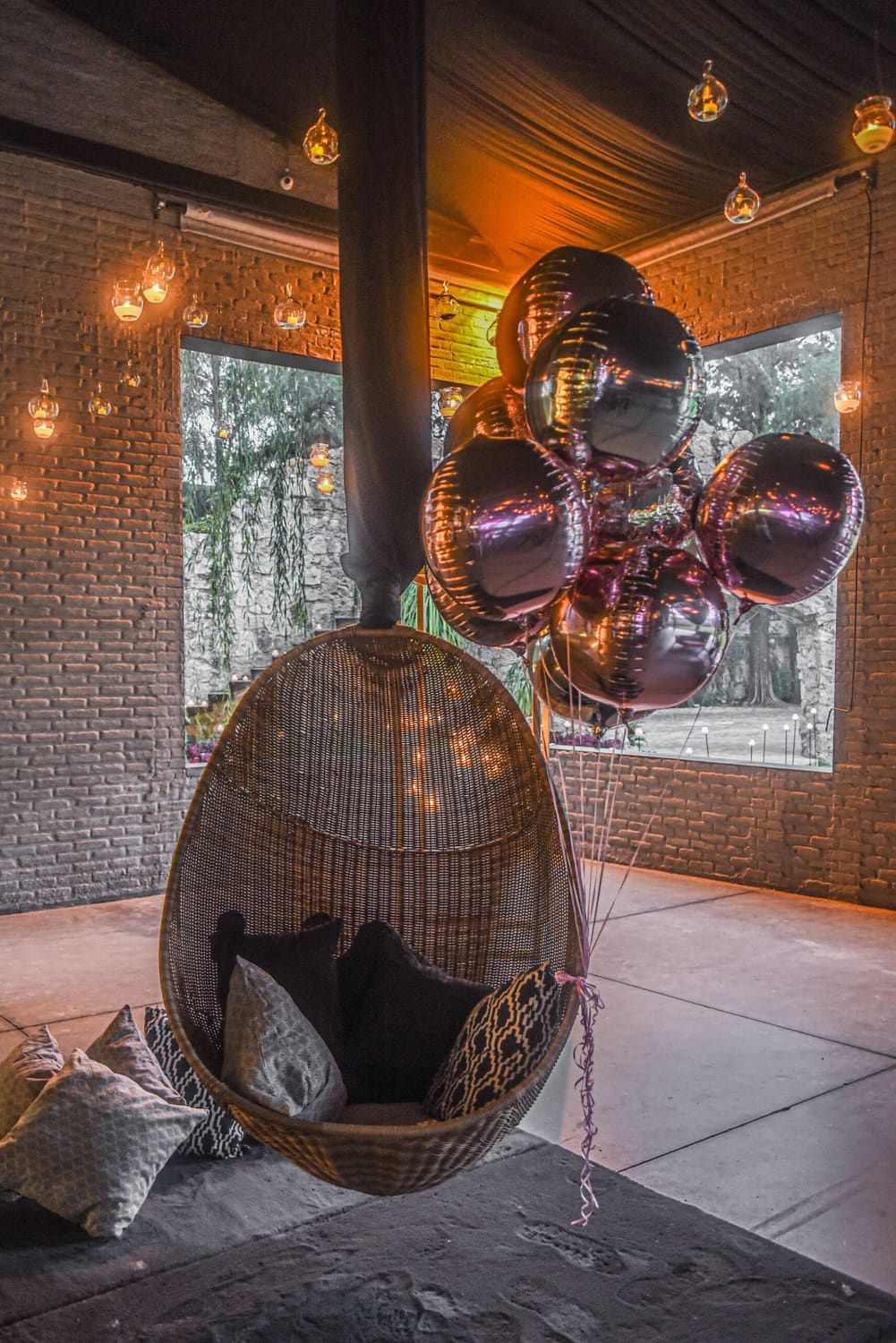 Sillón colgante de ratán con globos metálicos frente a un muro de ladrillo y luces cálidas