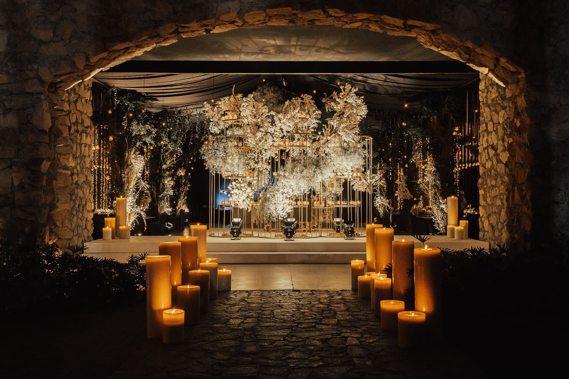 Instalación floral blanca bajo un arco de piedra, con columnas de velas encendidas a los lados