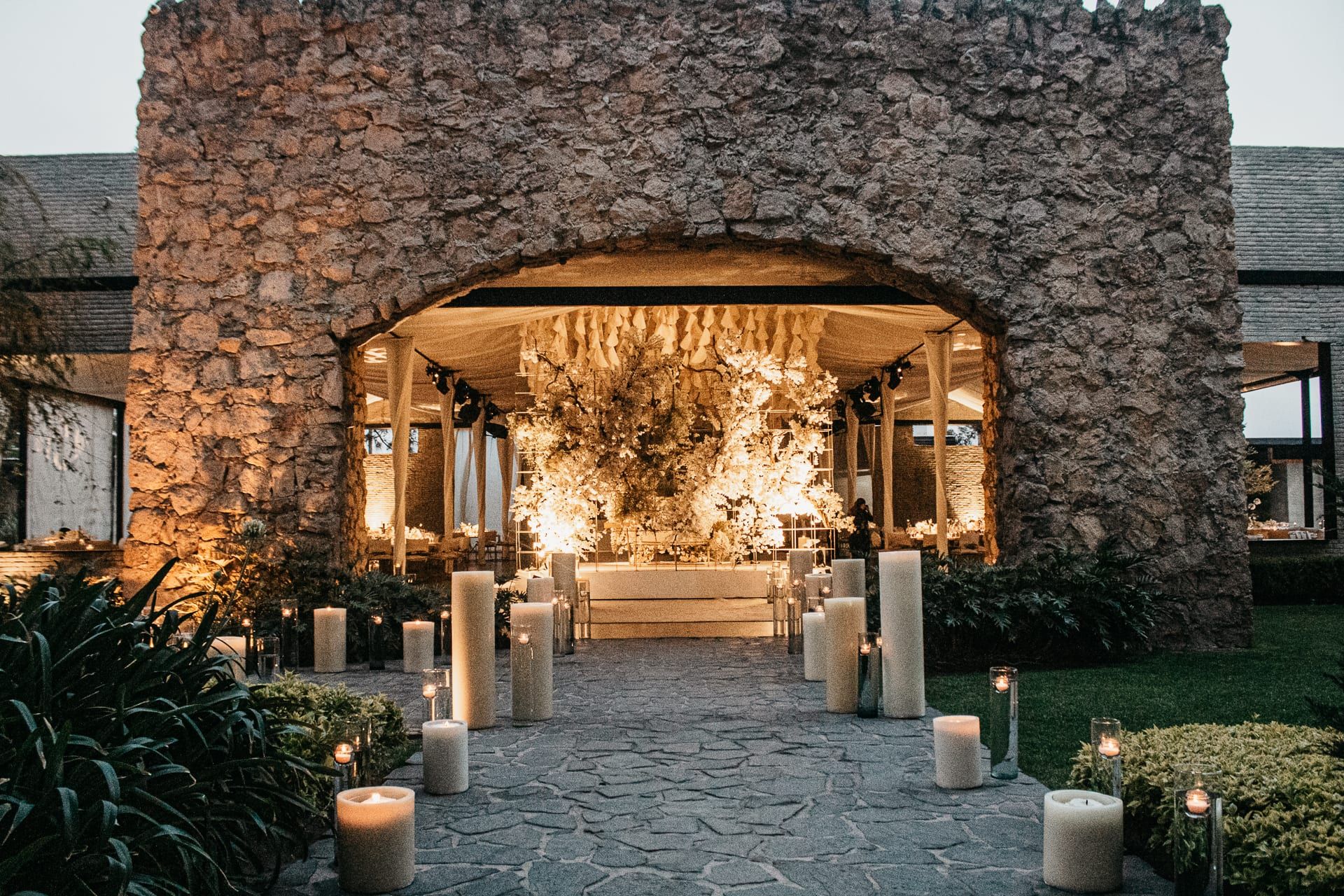 Entrada de piedra iluminada con un camino de velas y flores, y la recepción al fondo