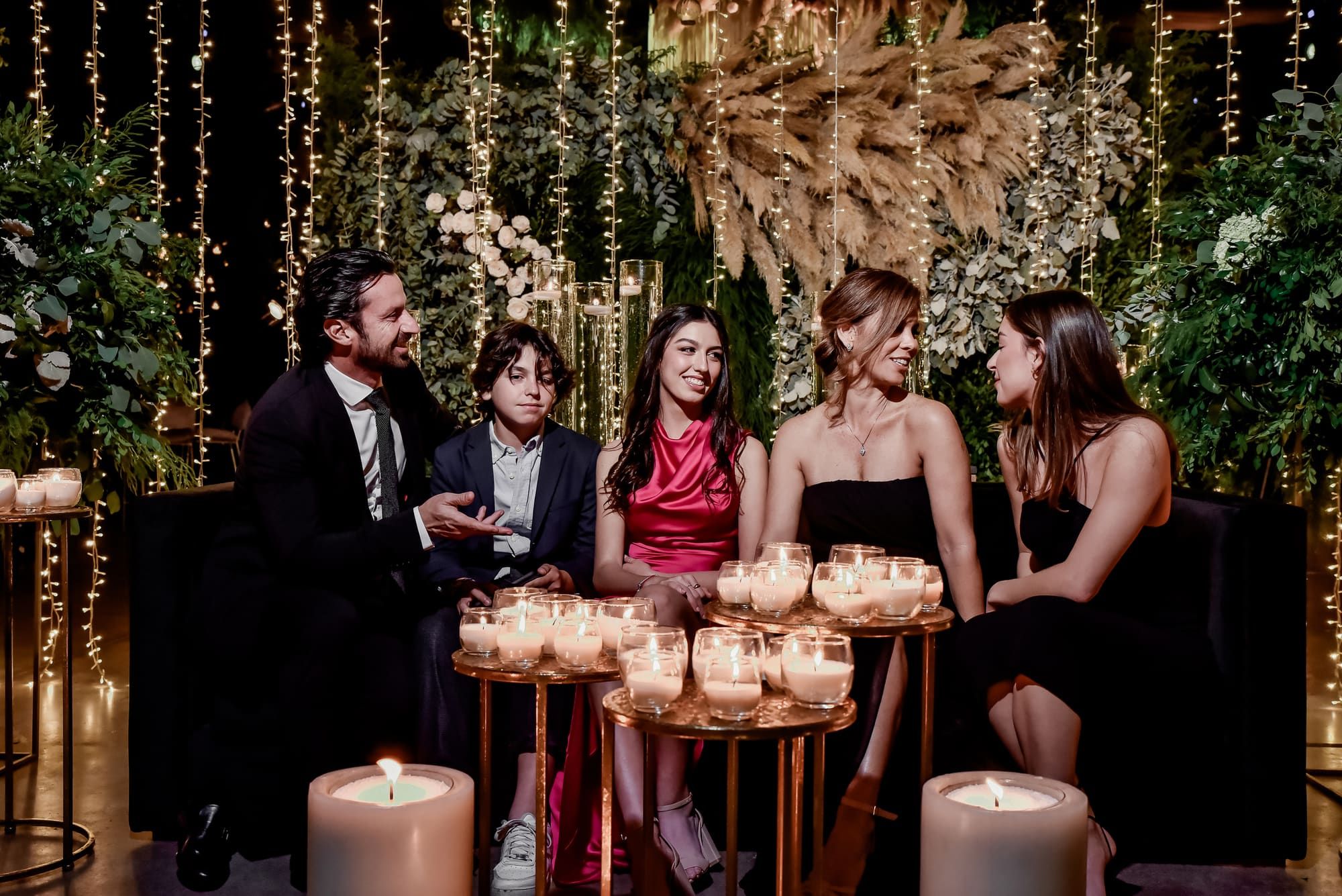 Cinco personas elegantes sentadas en un sillón negro ante mesas de cobre con velas, entre follaje y luces colgantes