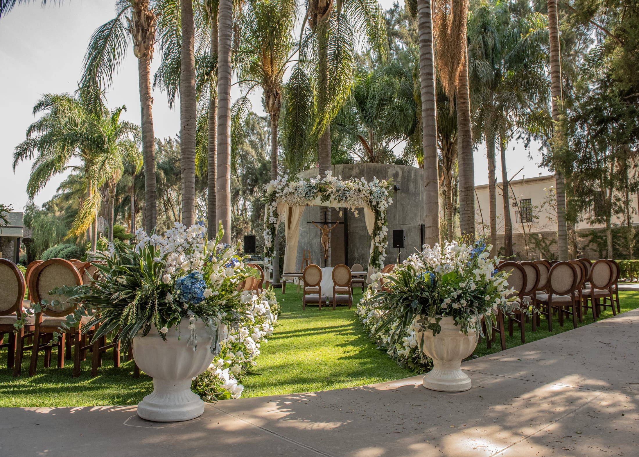 Dos grandes arreglos de flores blancas y azules en macetones sobre el césped, con una pérgola floral al fondo