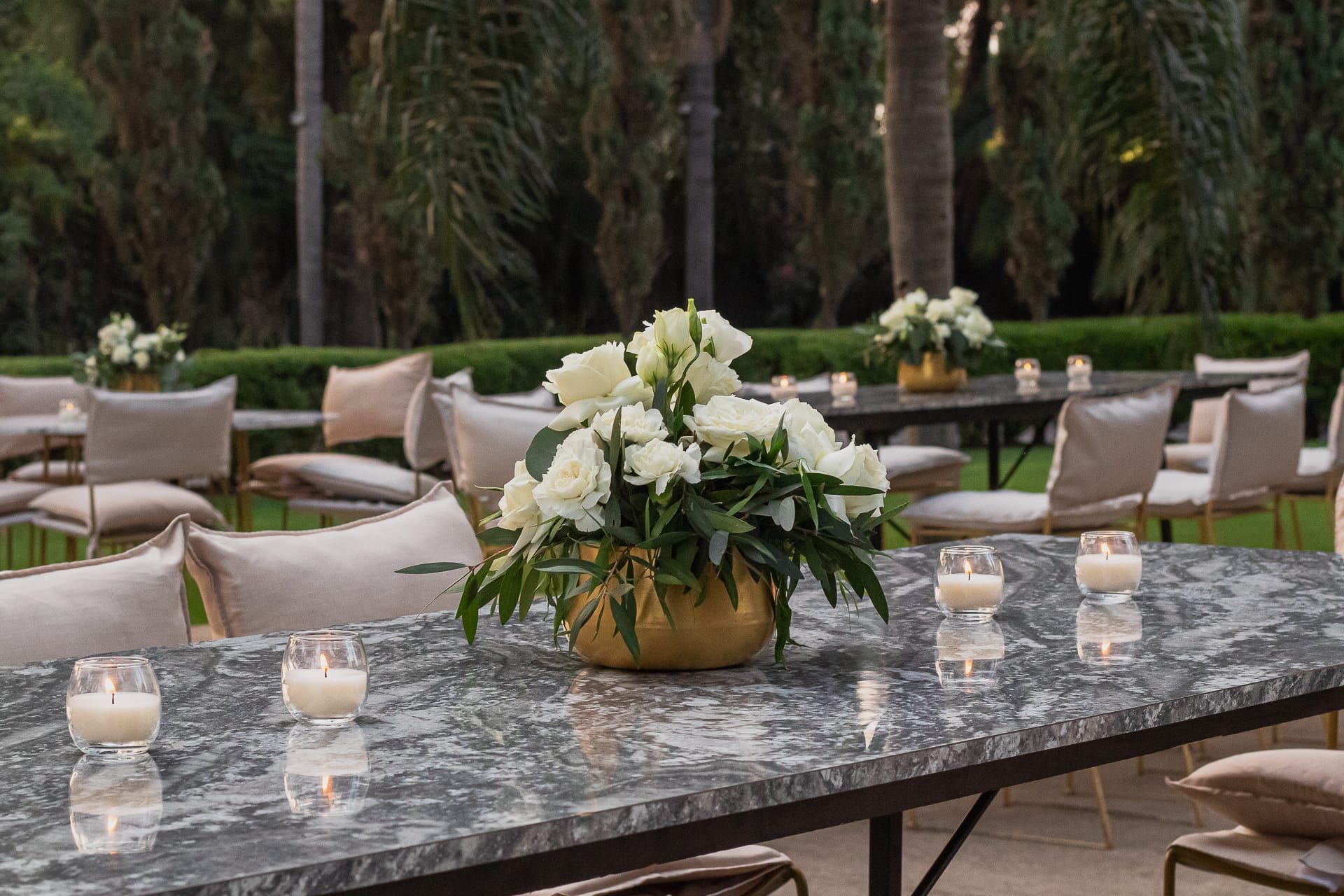 Mesa de mármol gris con arreglo de flores blancas en un florero dorado y velas, en el jardín