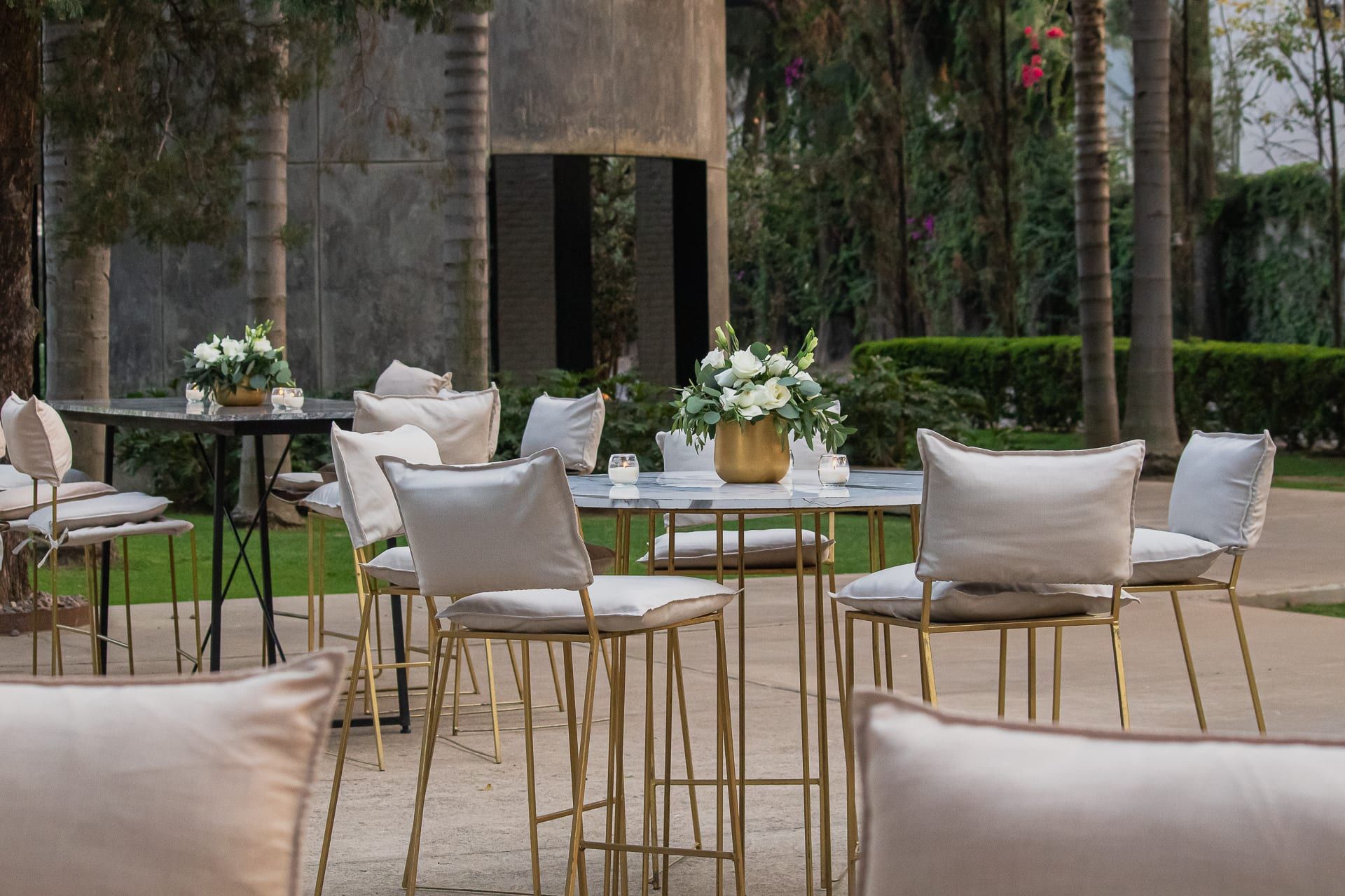 Mesas altas con taburetes dorados y sillones claros en el jardín, con arreglos blancos