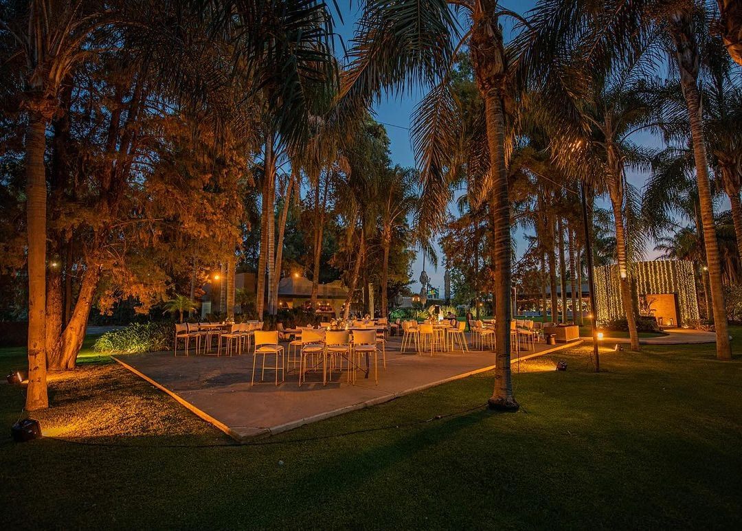 Jardín iluminado de noche con mesas y sillas entre palmeras