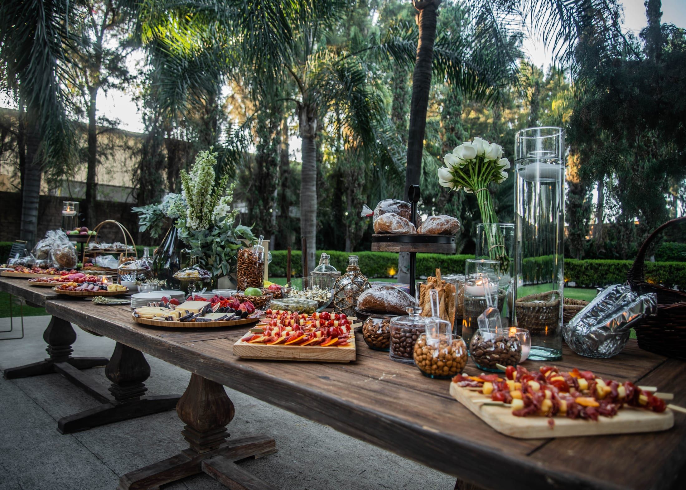 Larga mesa de madera con tablas de botanas, frutas y dulces al aire libre, entre palmeras