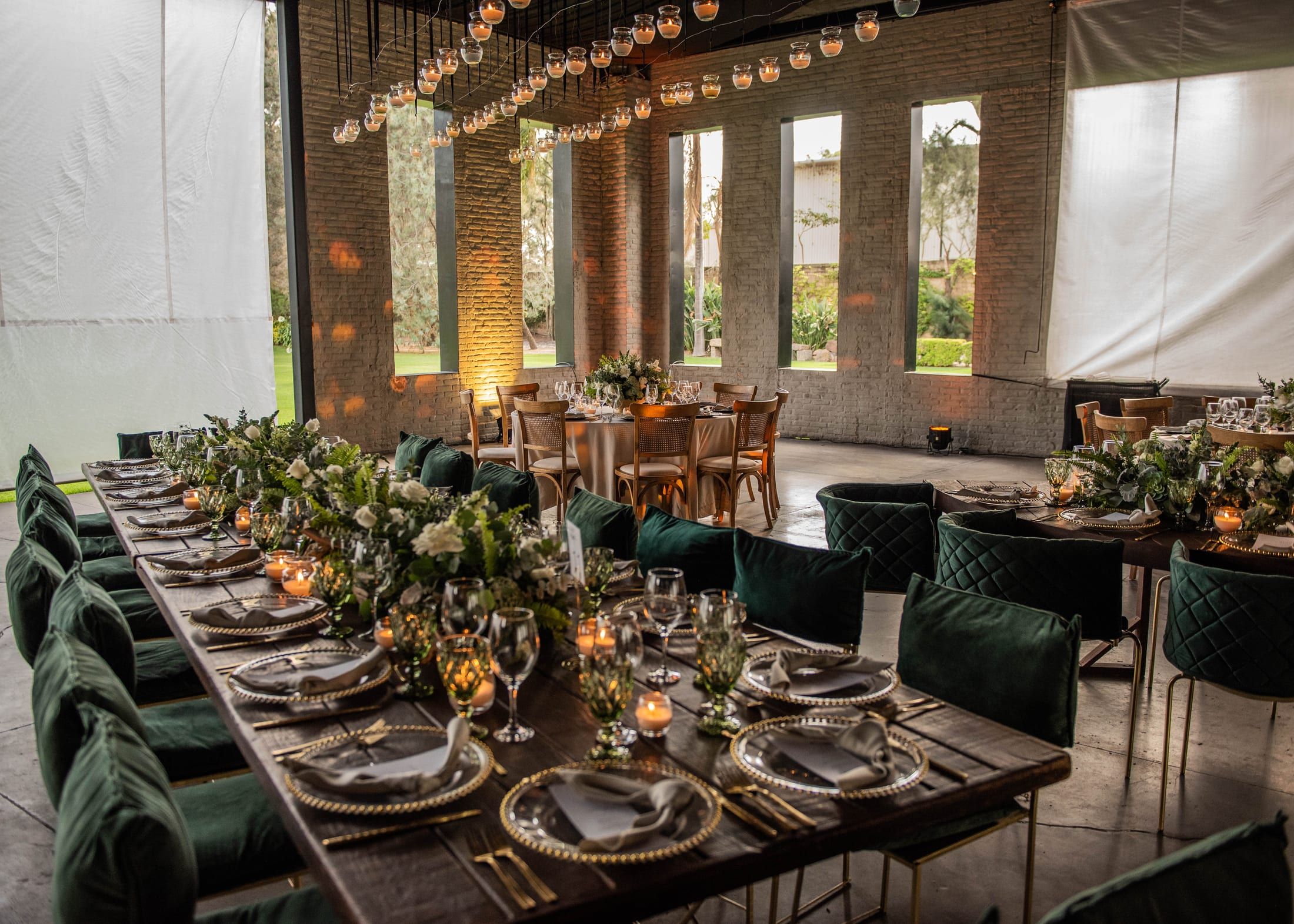 Larga mesa de banquete con sillas verdes acolchadas y centros de flores y velas, junto a ventanales