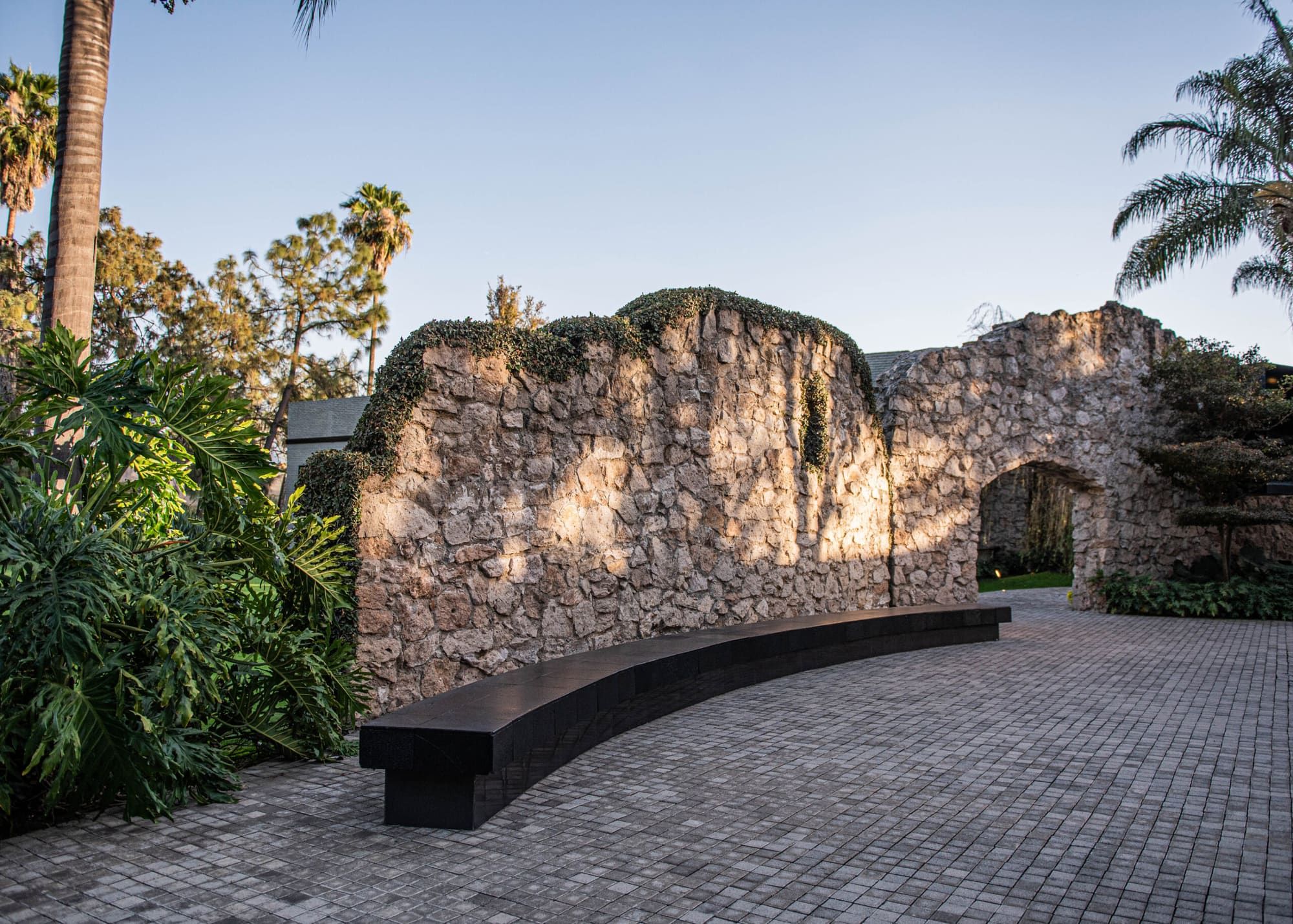 Muro de piedra con un arco y una banca curva oscura sobre un patio empedrado