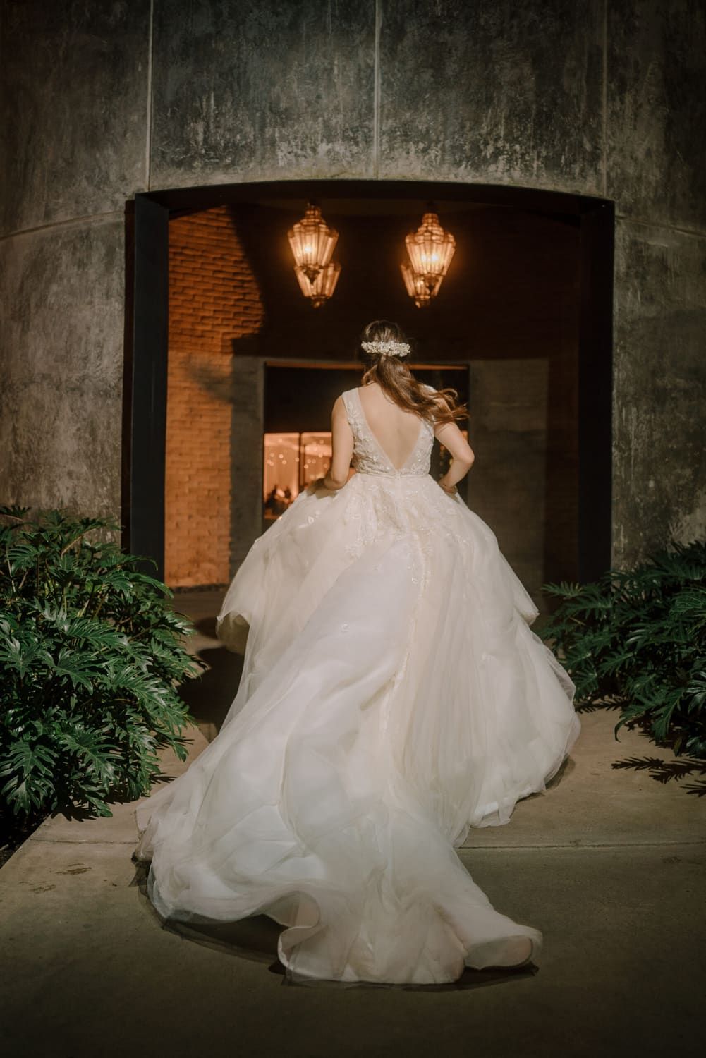 Novia de espaldas con vestido de falda amplia caminando de noche hacia una entrada de concreto con faroles