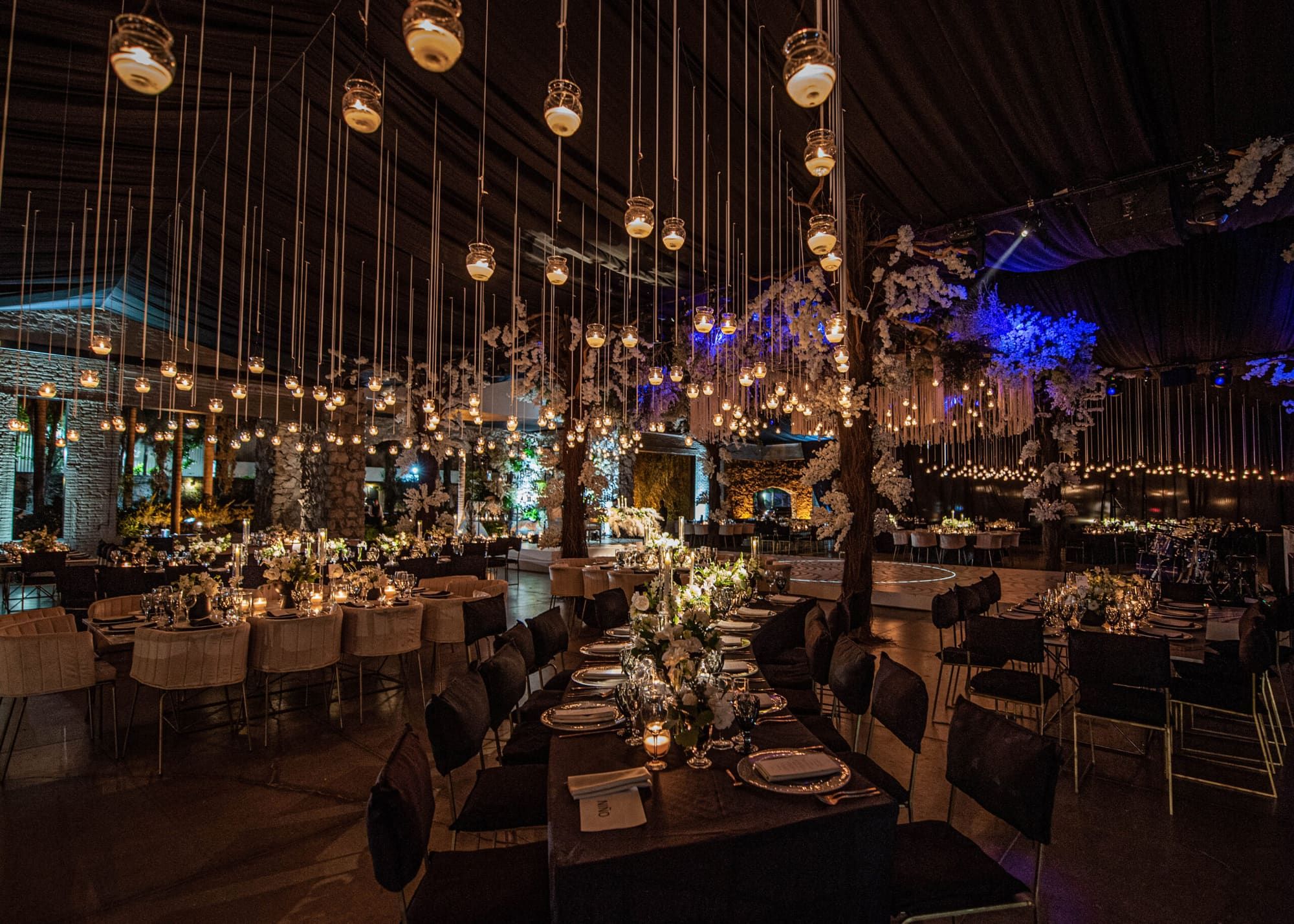 Salón de noche con faroles colgantes, árboles con flores blancas, luz azul al fondo y mesas de banquete