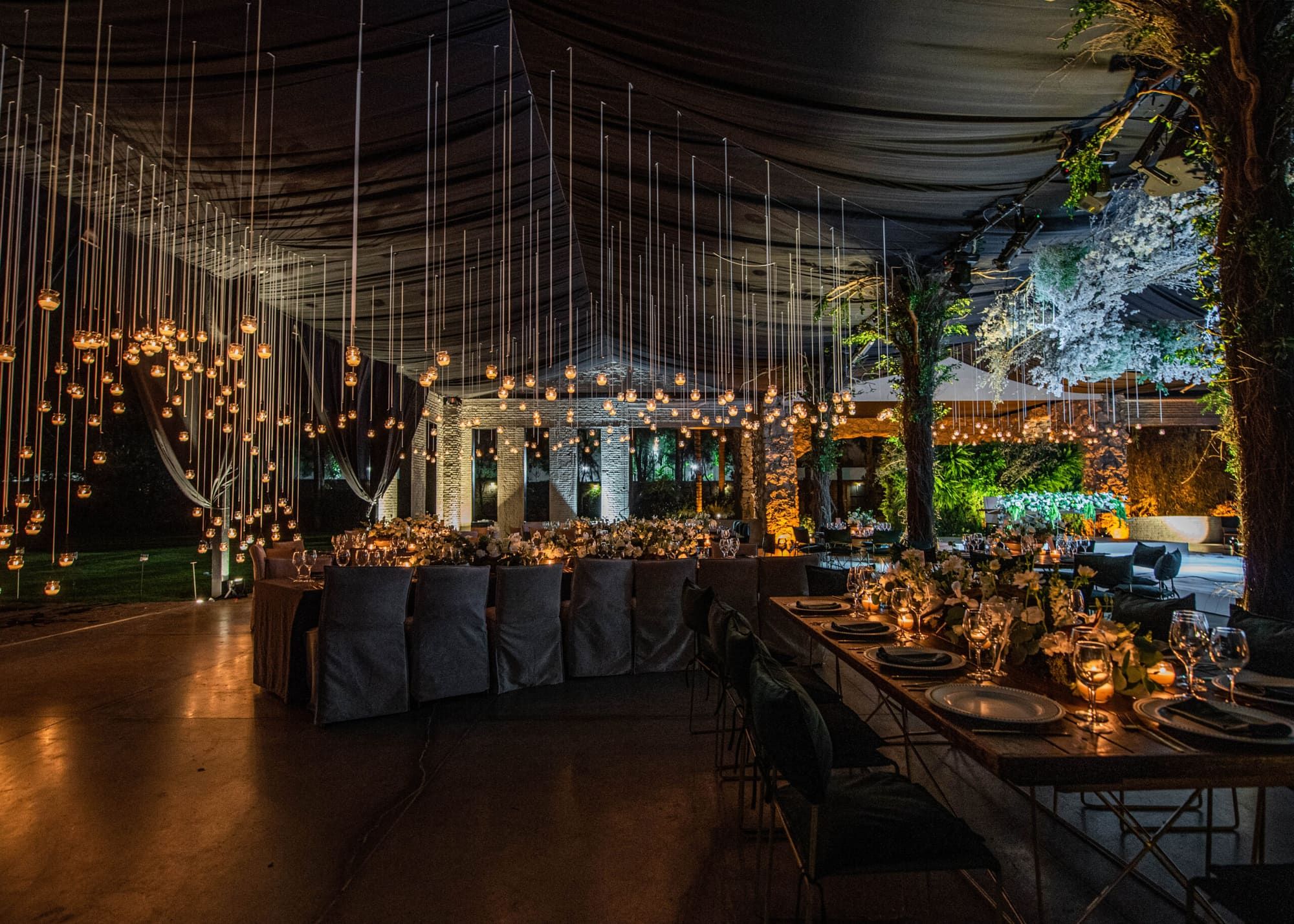 Salón de noche con larga mesa de banquete, hilos de luces colgantes, techo de telas oscuras y un árbol floral