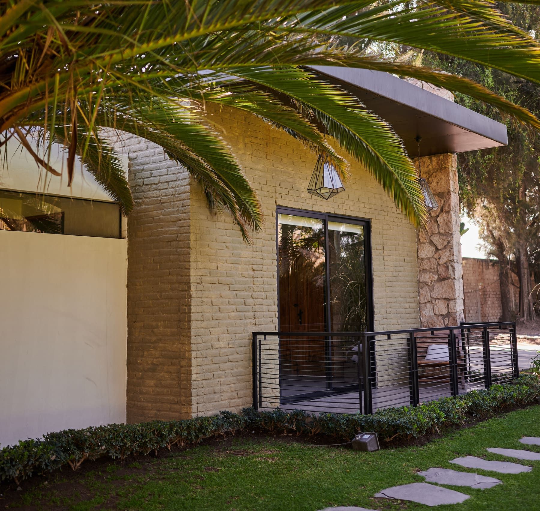Fachada de ladrillo y piedra con puerta corrediza de cristal, terraza con barandal y hojas de palmera