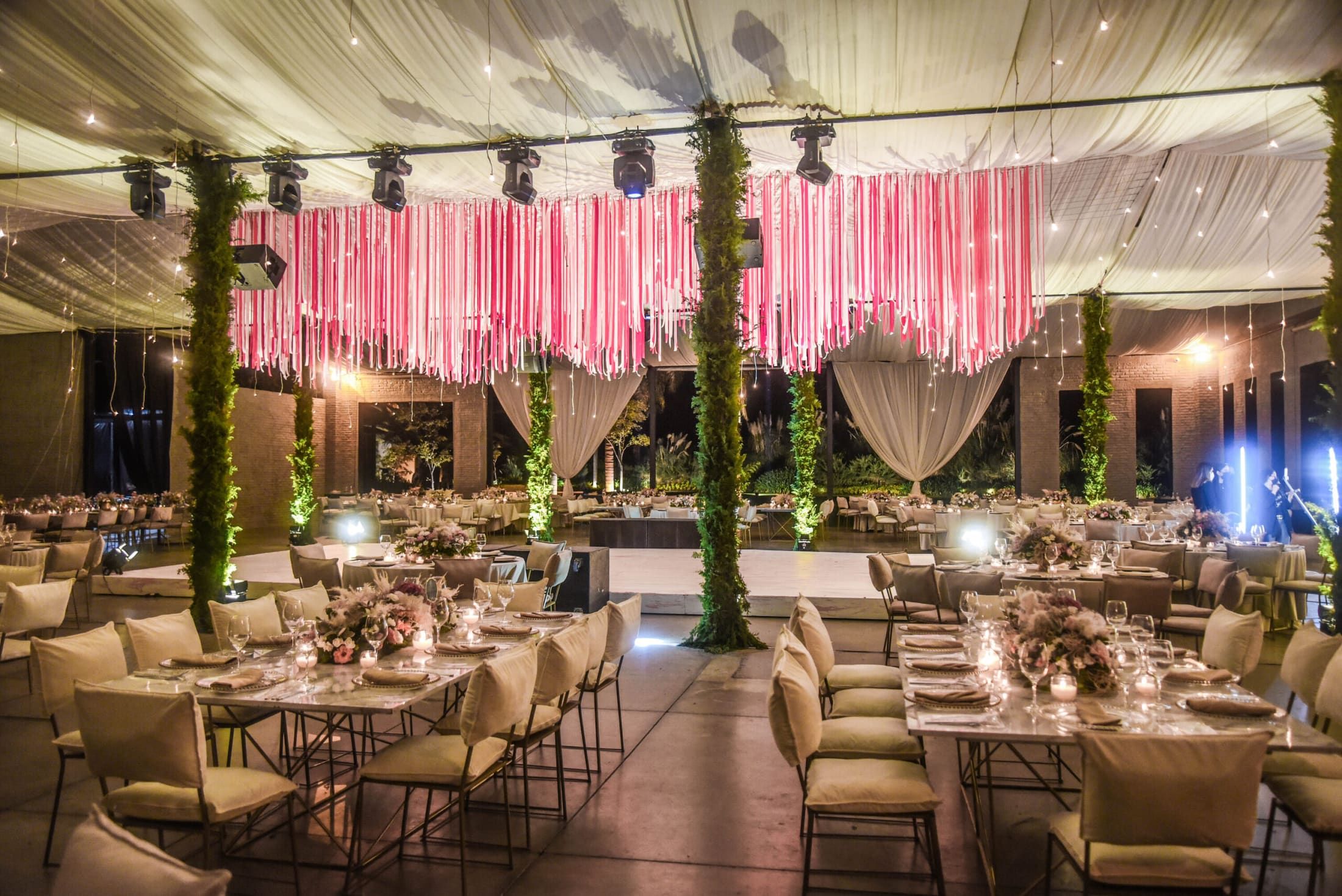 Salón con techo de telas blancas, cortina de listones rosas y mesas de banquete con sillas claras