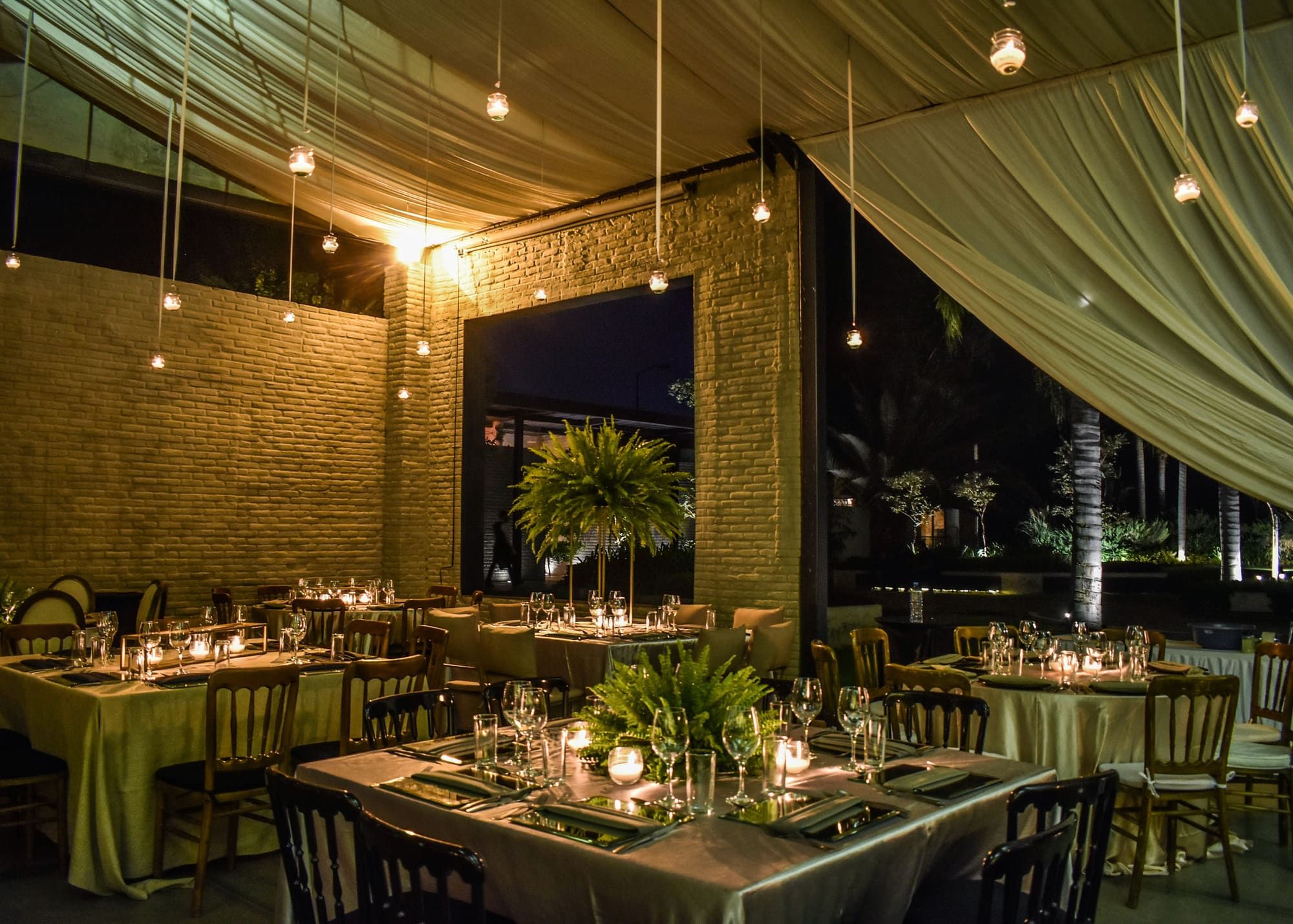 Salón con techo de telas, muros de ladrillo y mesas montadas bajo luces cálidas colgantes
