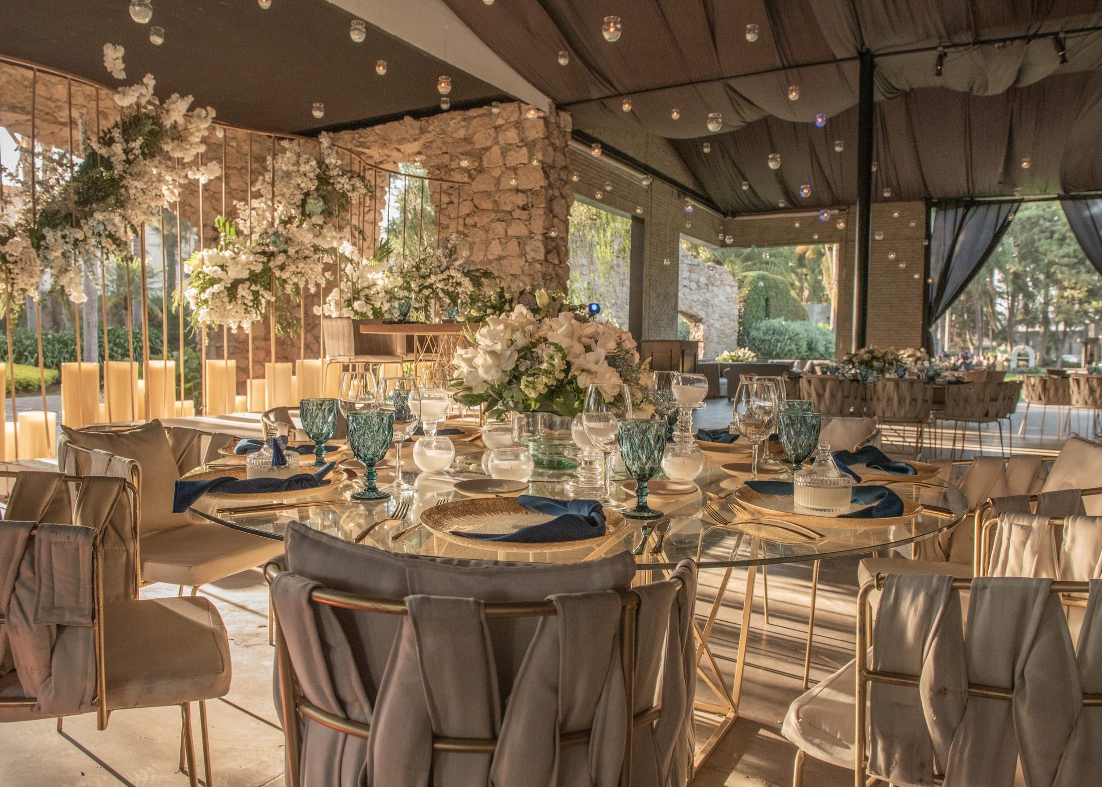 Mesa de banquete con copas azules y arreglos florales en el salón, con luz cálida y muros de piedra
