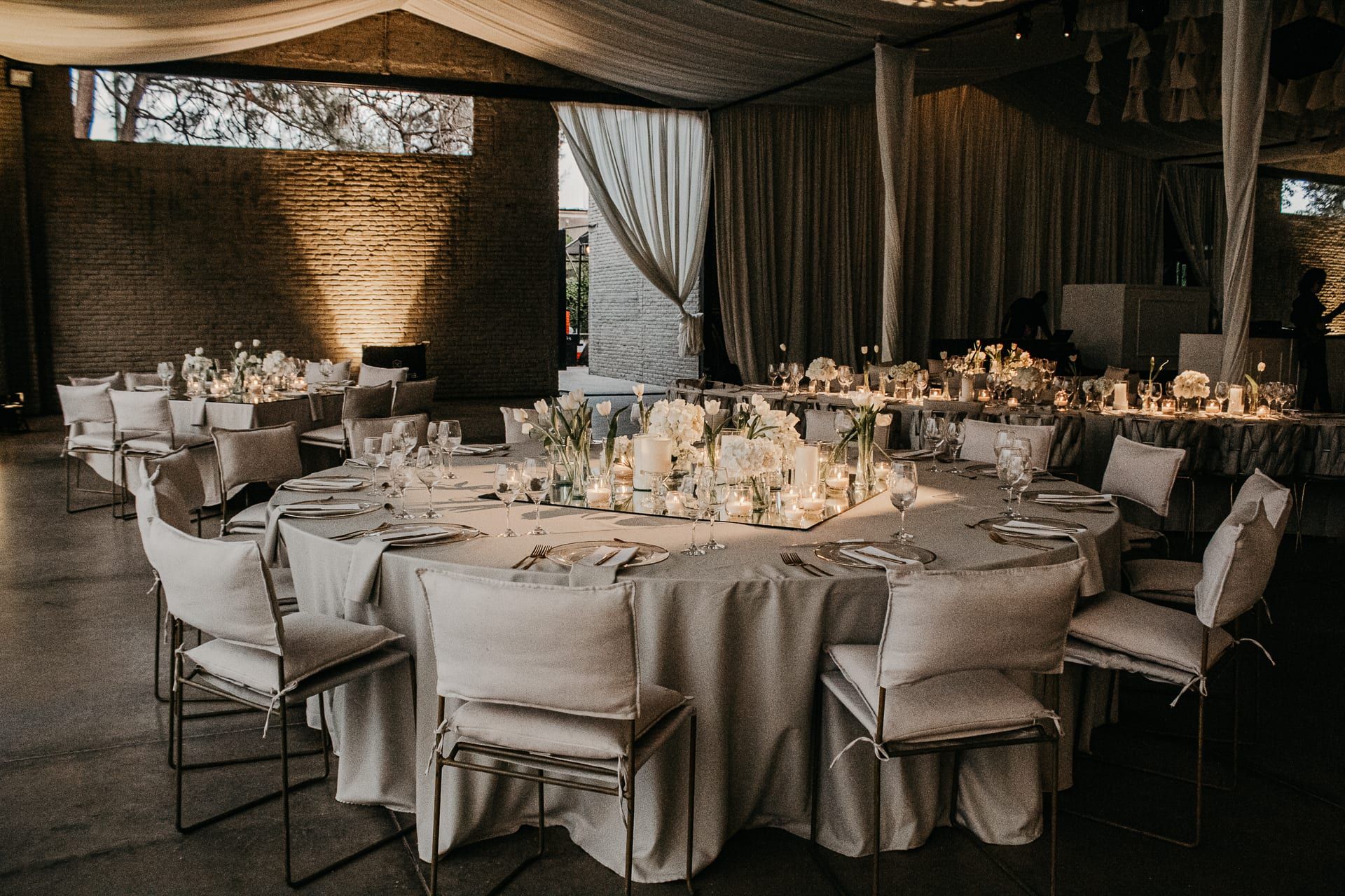 Salón con mesas redondas vestidas de lino claro, sillas blancas y cortinas, bajo luz cálida