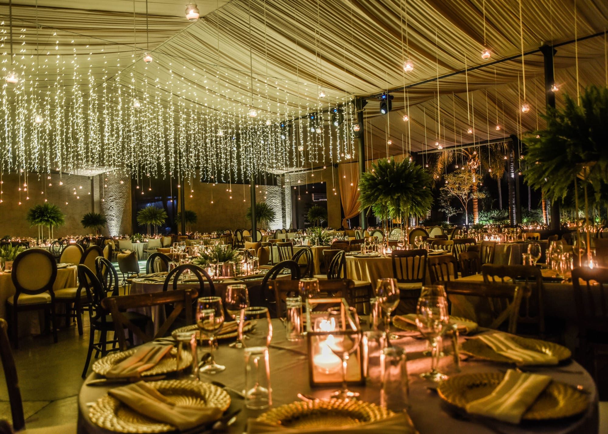Mesa redonda con platos base dorados, copas y farol con vela, bajo cortinas de luces