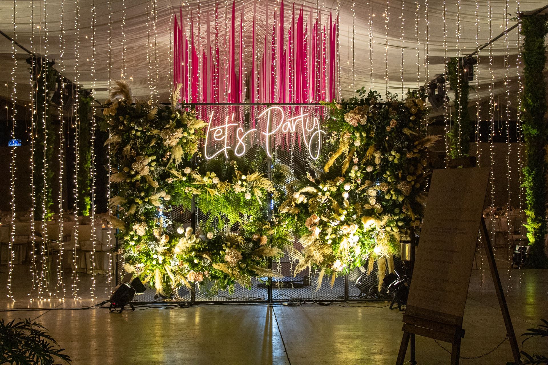 Muro de follaje y flores con letrero de neón «Let's Party» y listones rosas bajo cortinas de luces