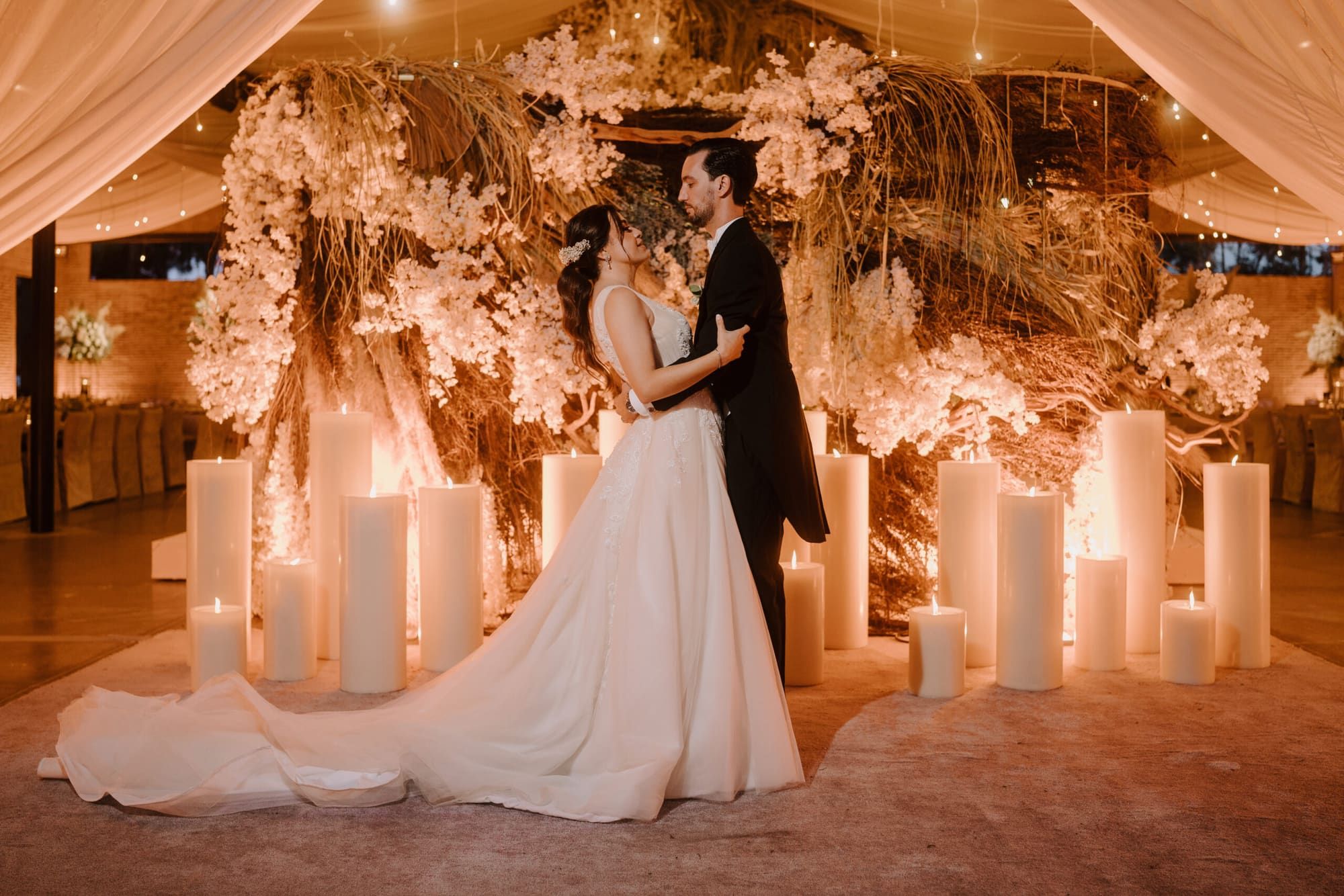 Pareja de novios abrazados entre columnas de velas y arreglos florales blancos, bajo cortinas de tela