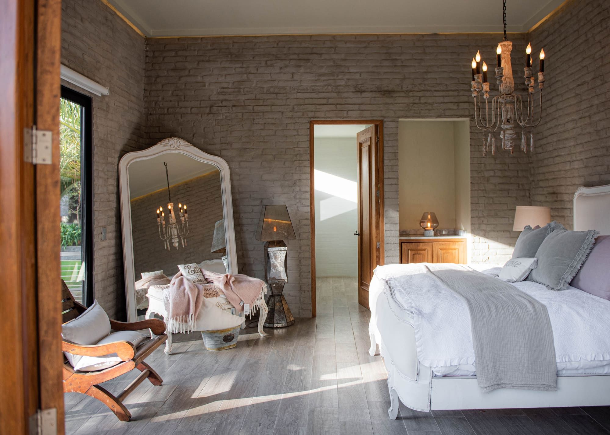 Suite con cama blanca, espejo de marco blanco, candil y luz natural entre muros de ladrillo