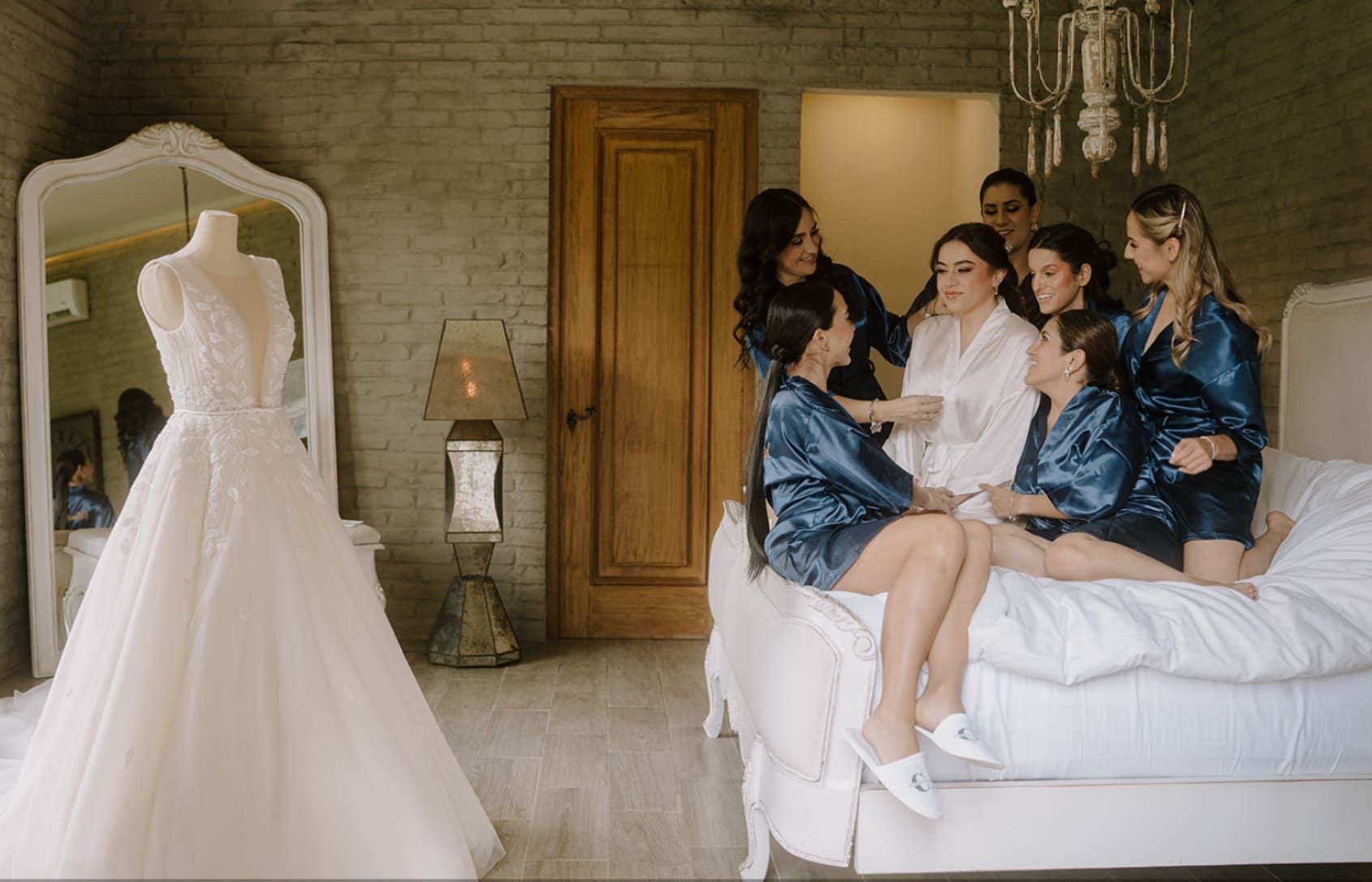 Mujeres con batas satinadas sentadas sobre la cama, junto a un vestido de novia en un maniquí