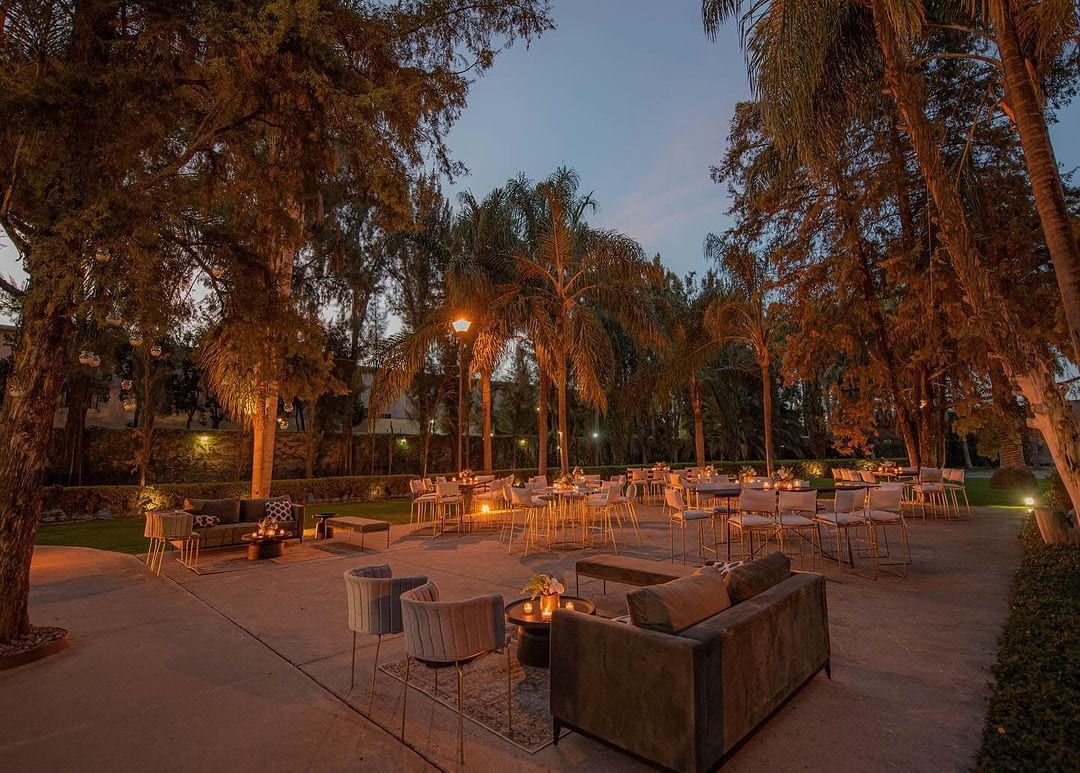Terraza de noche con sillones, mesas altas y luz cálida entre palmeras